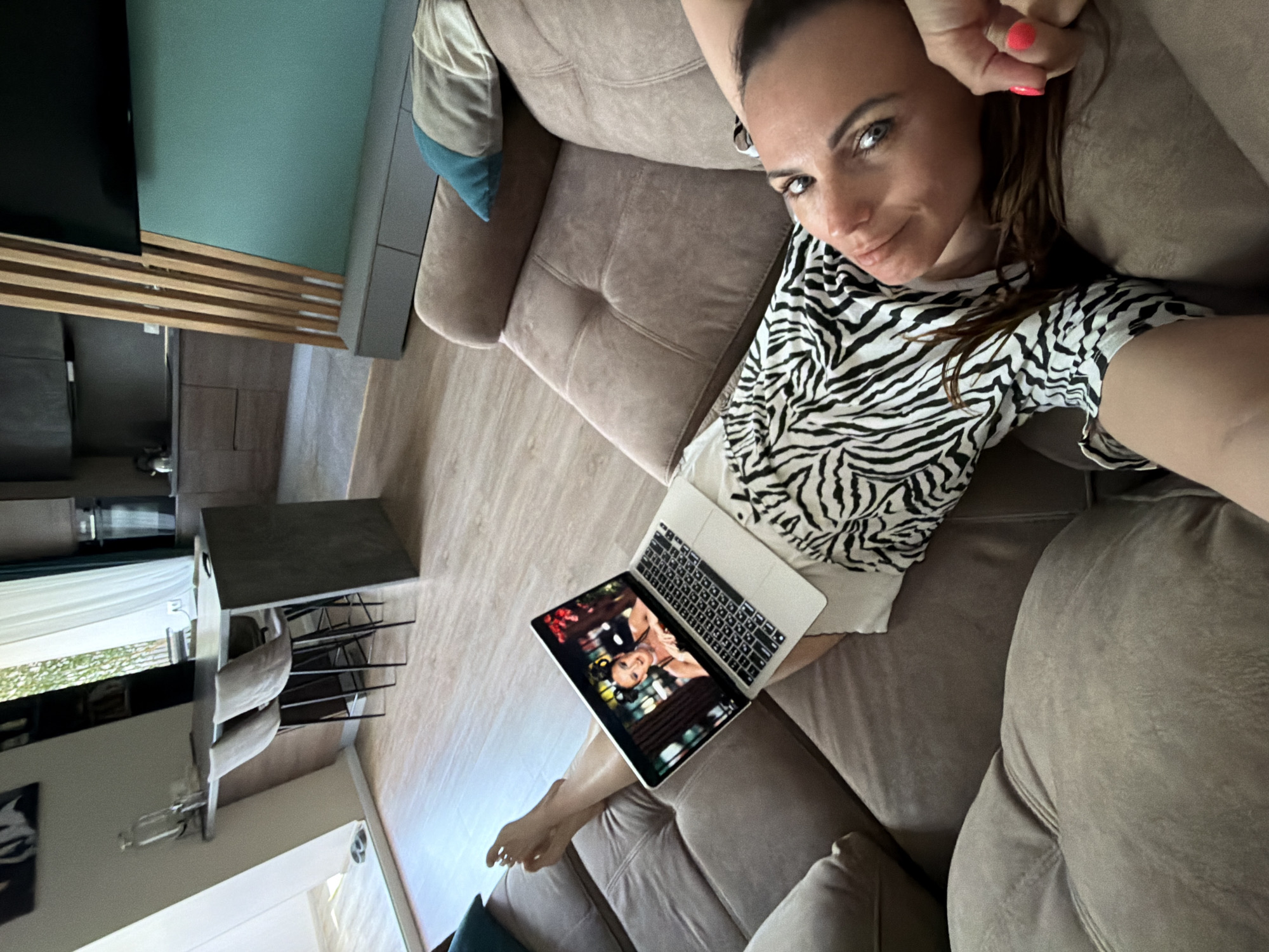 A woman is reclining on a brown couch indoors, holding a laptop open on her lap. She is wearing a zebra-print shirt and white shorts. She is looking at the camera and has her right hand near her head. The laptop screen shows an image of two people. A blue pillow is on the left side of the couch. A wooden TV stand is visible in the background, along with a dining table set. The scene is not located in the city of Birobidzhan, Russia.