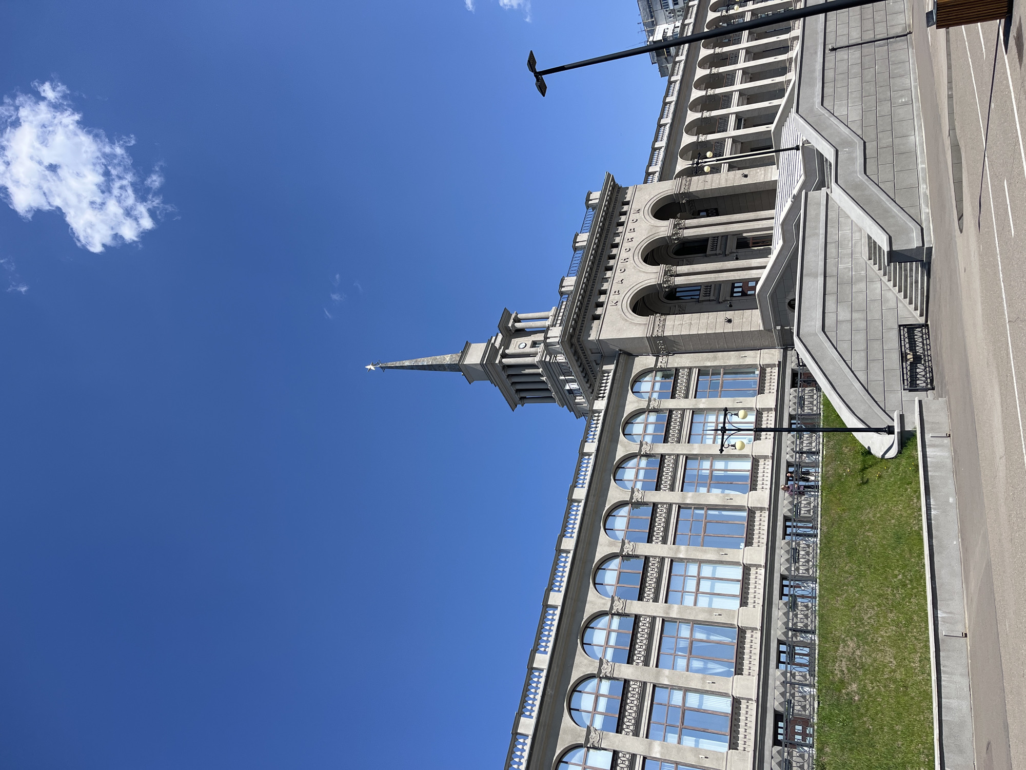 The image shows a building in Krasnoyarsk, Russia. The building is made of gray stone and has a tall spire with a star on top. The building has arched windows and columns. A wide staircase leads to a higher level of the building. There is a green lawn between the building and a road. A lamppost stands in front of the building. A patch of white cloud is visible in the upper left corner of the image. The sky is clear and blue.