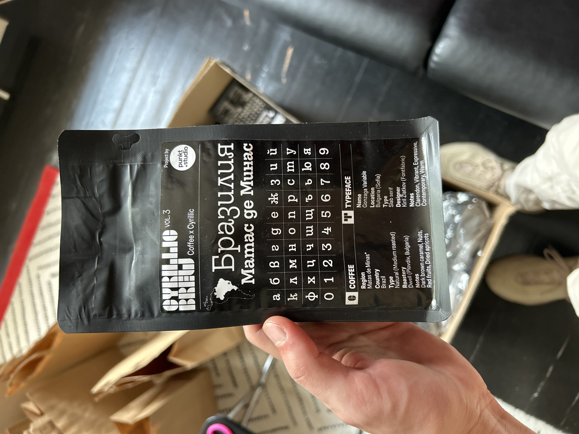 The image shows a person holding a black coffee package with Cyrillic lettering. The package is for "Brazil Matas de Minas" coffee and includes information in English, such as the coffee type, roastery (Dwell, located in Plovdiv, Bulgaria), and tasting notes. The package also details the typeface used, "Gonzaga Variable," its location as Bulgaria (Sofia), its type as Slab serif, and its designer, Kiril Zlatkov (Fontfabric). The hand holding the package is visible against the background of an opened cardboard box. A blurred part of a sofa and floor are also visible in the background. The scene suggests someone is examining or presenting the coffee package, possibly in an indoor setting. The package itself is the primary object of interest.