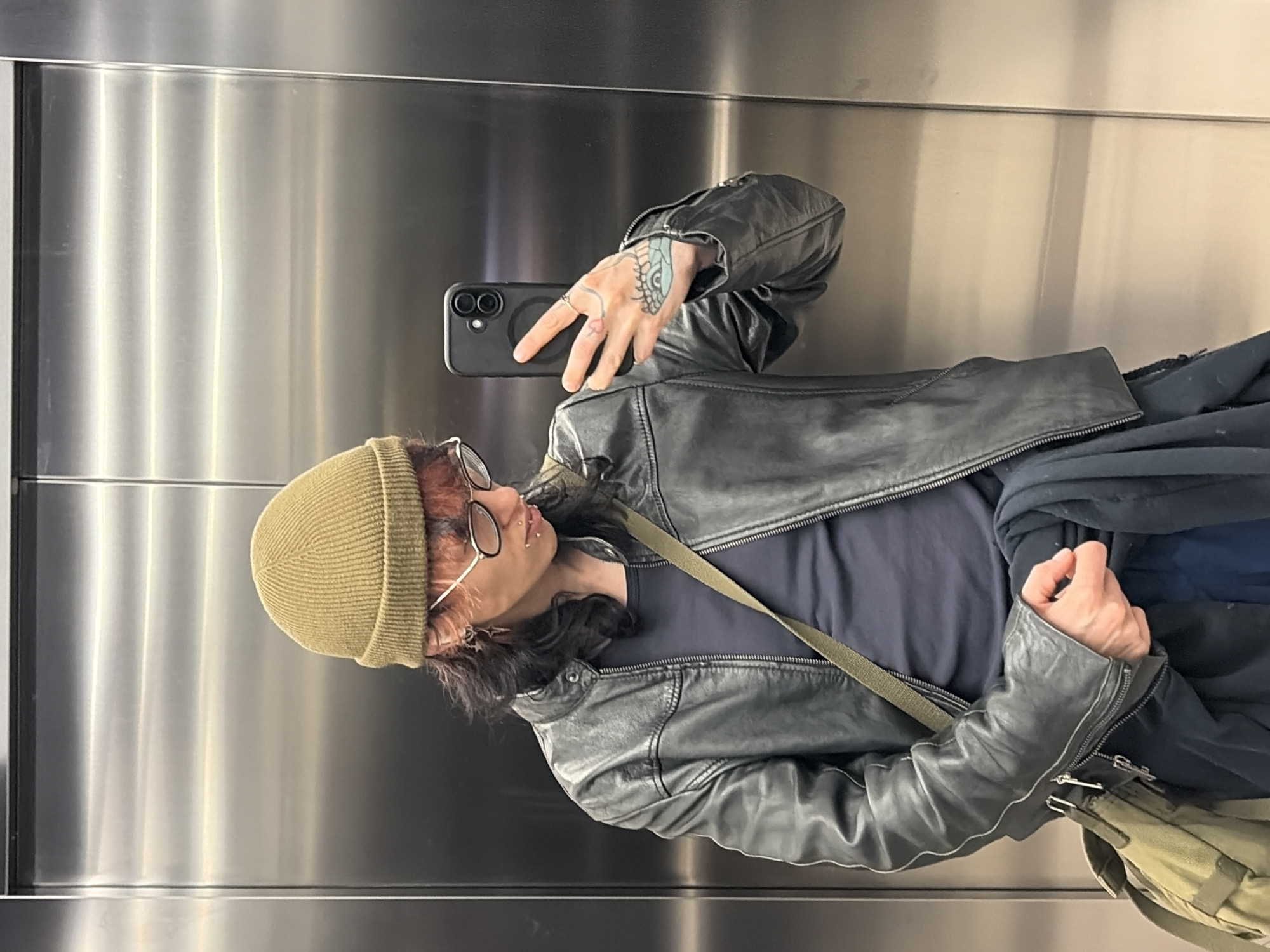 A person takes a selfie in an elevator. The person is wearing a green beanie, glasses, a black leather jacket, and a dark shirt. They have a tattoo on their hand and piercings in their face. They are holding a black smartphone in their left hand. The elevator has stainless steel walls. It is not possible to verify that the location is in the city of Busmantsi, Bulgaria from this image.