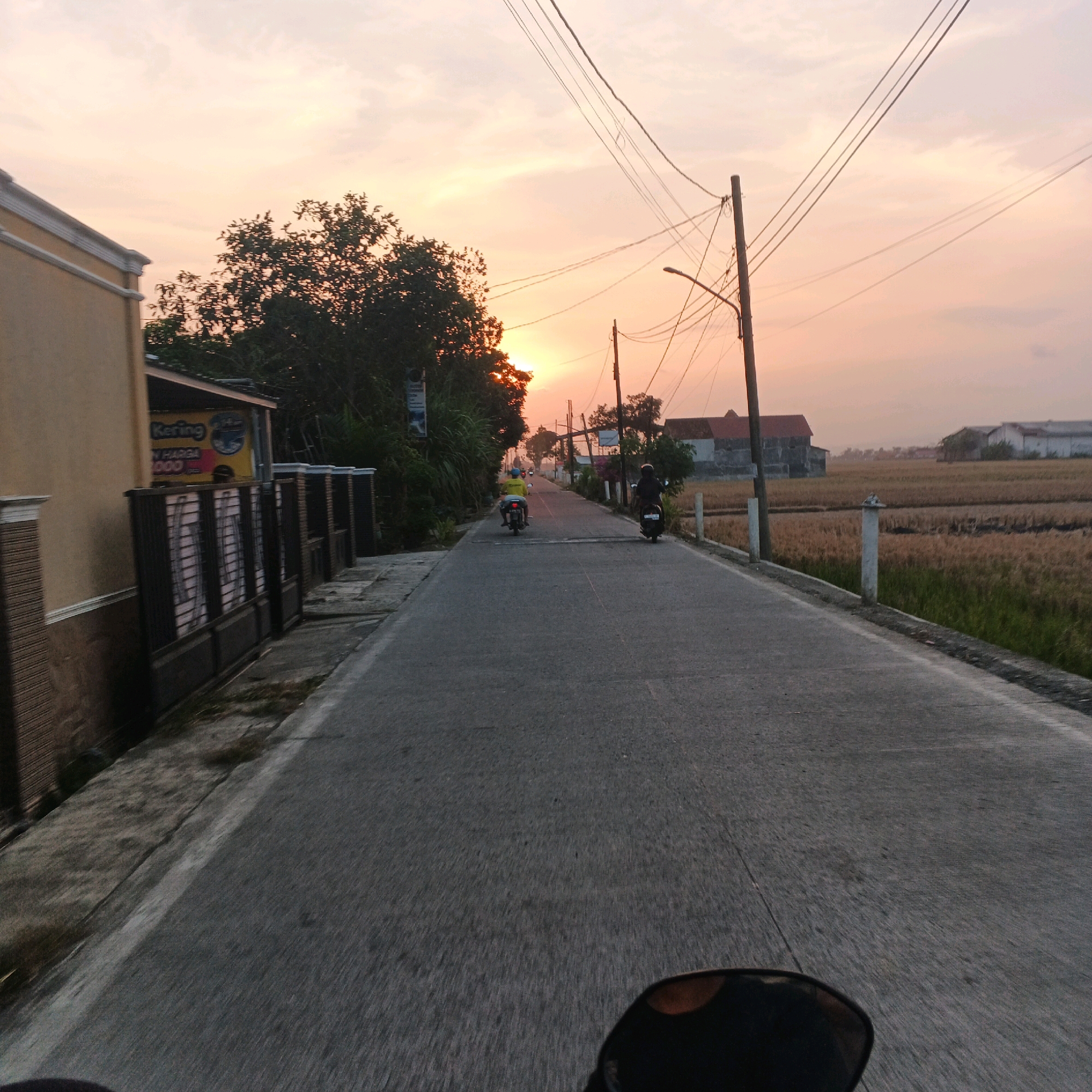 The image depicts a straight, paved road during sunset in Kalikudi, Indonesia. The sky is a soft orange and pink hue, with scattered clouds. Power lines stretch across the sky, supported by wooden utility poles. On the left side of the road, a pale yellow building with decorative trim and a dark fence with vertical slats is visible. A sign with blue and yellow lettering, partially obscured, is mounted on the building. On the right side of the road, a dry field of golden stalks, likely rice, stretches towards a cluster of buildings with red and white roofs in the distance. Two people on motorcycles are traveling away from the camera down the road. The first motorcycle, closer to the camera, has a rider wearing a dark helmet and clothing. The second motorcycle, further down the road, has a rider wearing a yellow jacket and a helmet. The perspective suggests the photo was taken from another moving motorcycle.