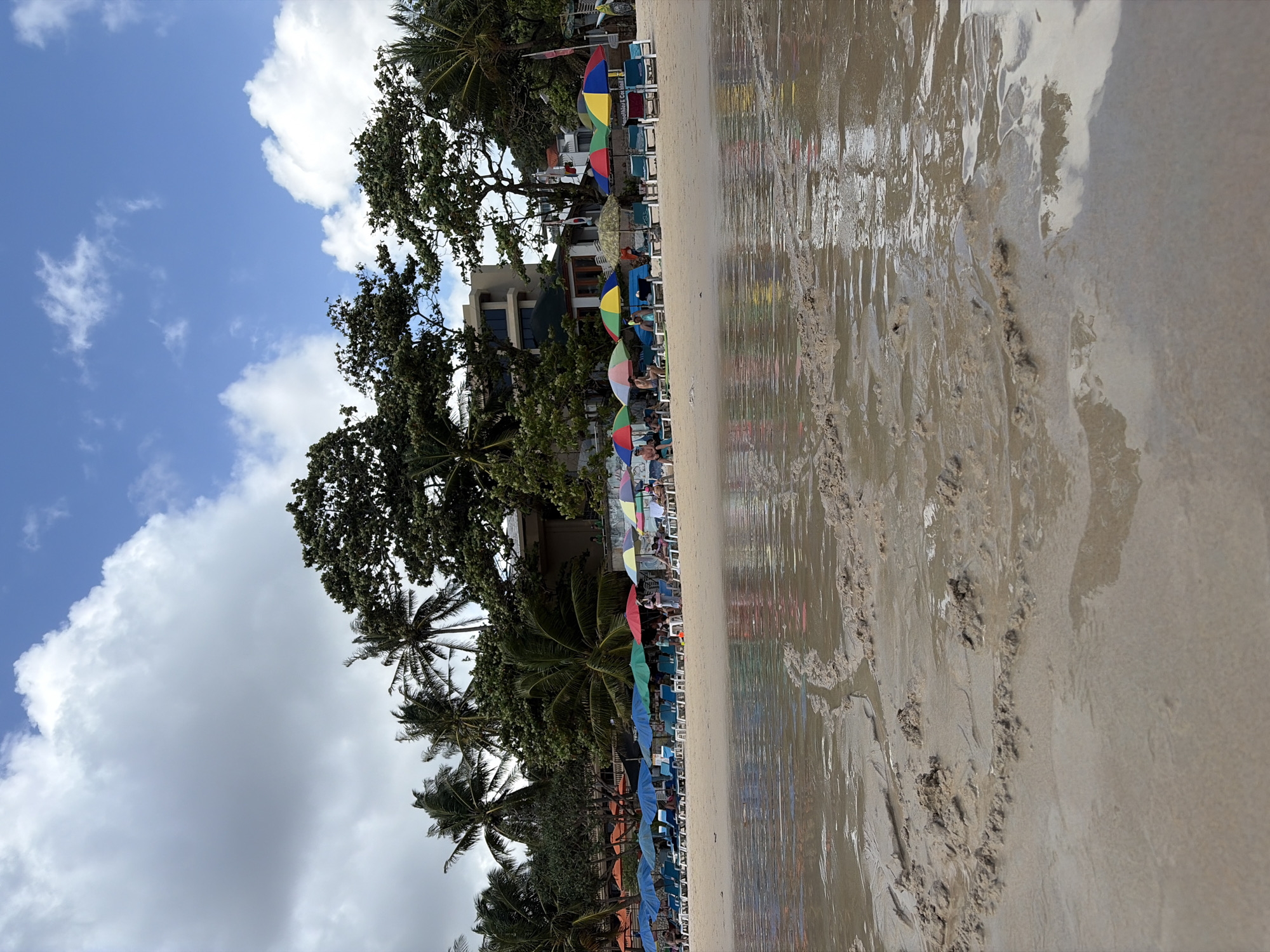 The image depicts a beach scene. In the foreground, wet sand with ripples and small mounds is visible, with shallow water covering parts of it. The middle ground features a row of lounge chairs and colorful beach umbrellas set up on the sand. Behind the chairs and umbrellas, lush green trees and a multi-story building can be seen. The sky above is blue with scattered white clouds. Several people are present, seated on the chairs or walking on the beach. The overall atmosphere suggests a sunny day at a resort beach.