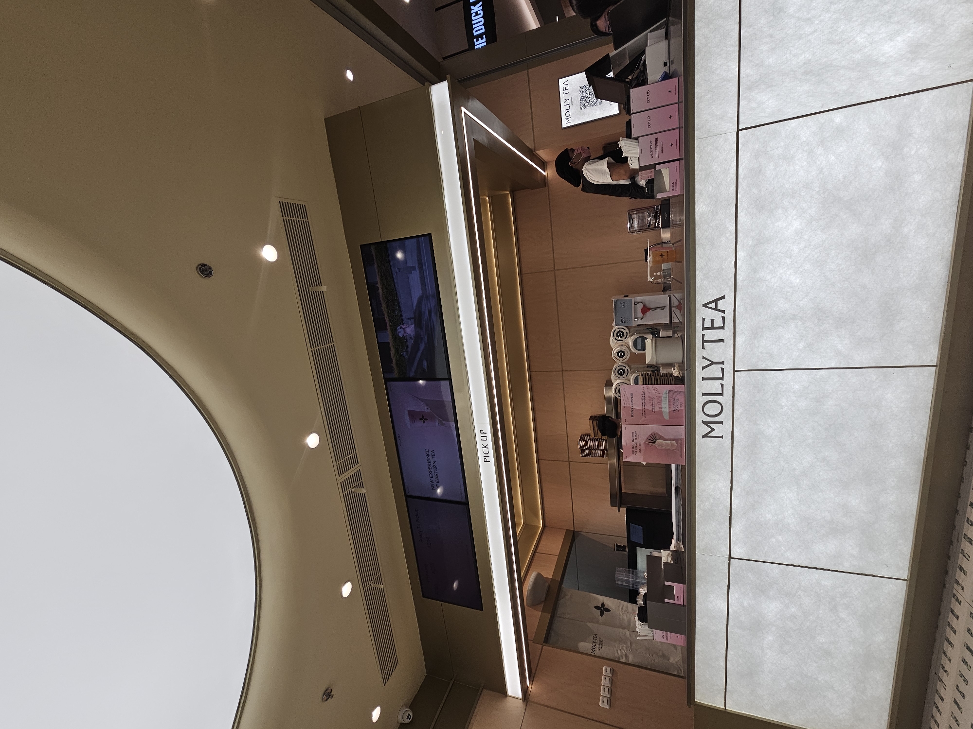 The image shows a modern tea shop called Molly Tea, likely located in a shopping mall or commercial area in Condongcatur, Indonesia. The shop features a bright, clean aesthetic with light-colored wood paneling and a white, illuminated counter. A digital display above the counter shows advertisements for "New Experience of Eastern Tea" and a "Ready to Pickup" notification.
At the counter, a female employee wearing a dark vest over a white shirt and a dark bandana is visible, looking at a computer screen. Various tea-making equipment and supplies, such as cups, lids, and a dispenser, are arranged on the counter. Several customers are present, though their faces are not clearly visible. The floor in front of the counter has a patterned design. The overall environment suggests a busy but well-organized service establishment.