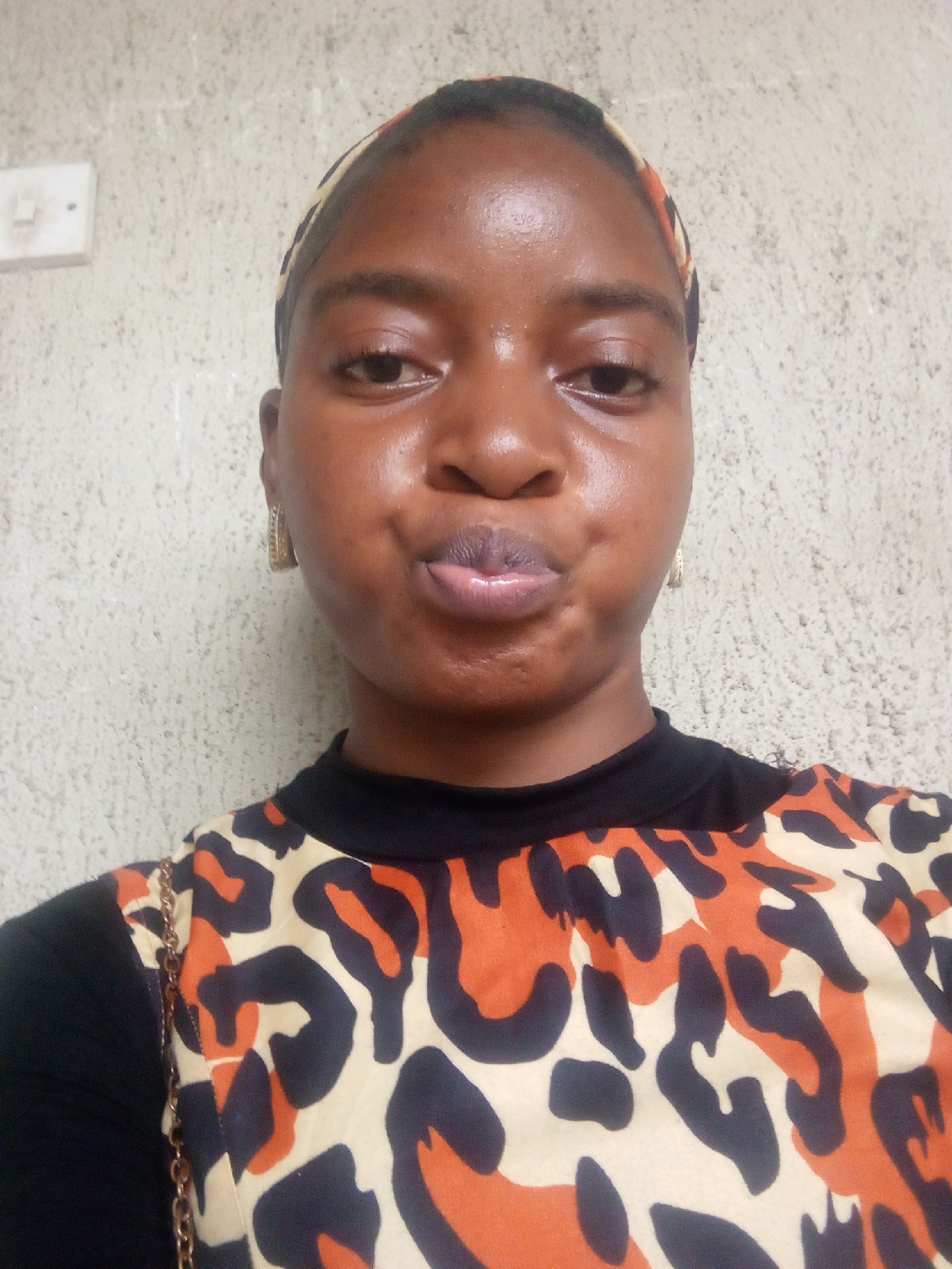 A young Black woman, likely in her late teens or early twenties, poses for a selfie against a textured, light-colored wall. She has dark skin, dark eyes, and dark hair, which is partially covered by a patterned headband. She is wearing a black shirt with a leopard print overlay on the front, featuring black, orange, and cream colors. The woman is looking directly at the camera with her lips pursed, and she is wearing a pair of gold hoop earrings. To the left of the woman, on the wall, is a white electrical outlet. The image appears to have been taken indoors, with the lighting originating from the front, causing some shine on her forehead and nose. The location is stated to be Serti, Nigeria.