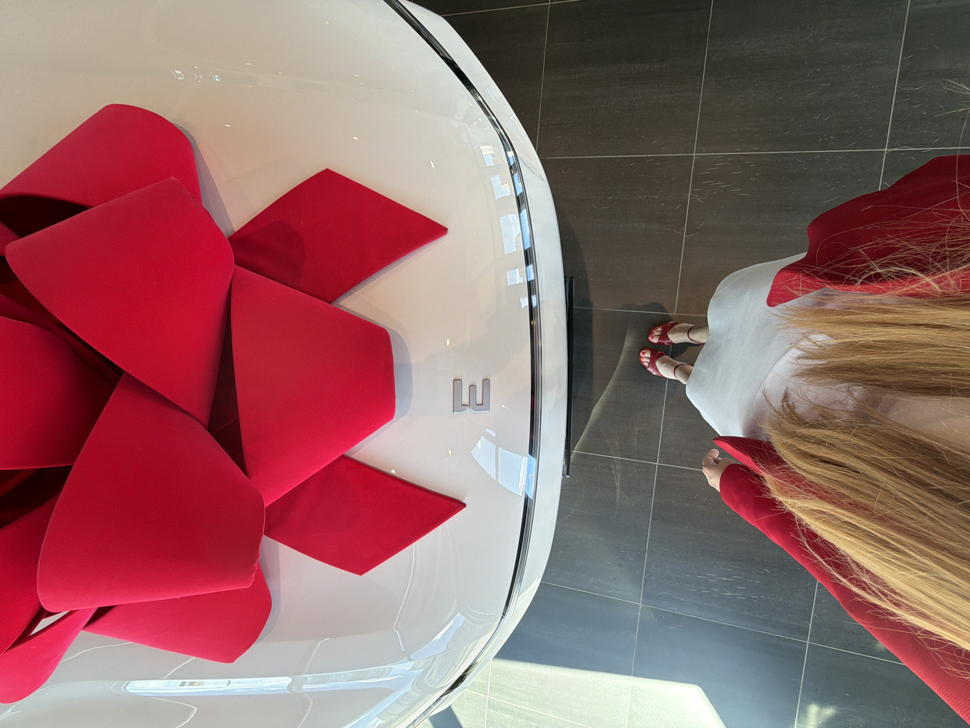 The image shows a white car with a large red bow on its hood. A person's feet, wearing red sandals, are visible at the bottom of the frame, along with the back of their head, showing long blonde hair and a red jacket. The car appears to be in a showroom, with tiled flooring. The scene is brightly lit, with reflections visible on the car's surface. The logo "E" is visible on the car. The image does not contain any information that would allow for determining its location in Izhevsk, Russia.