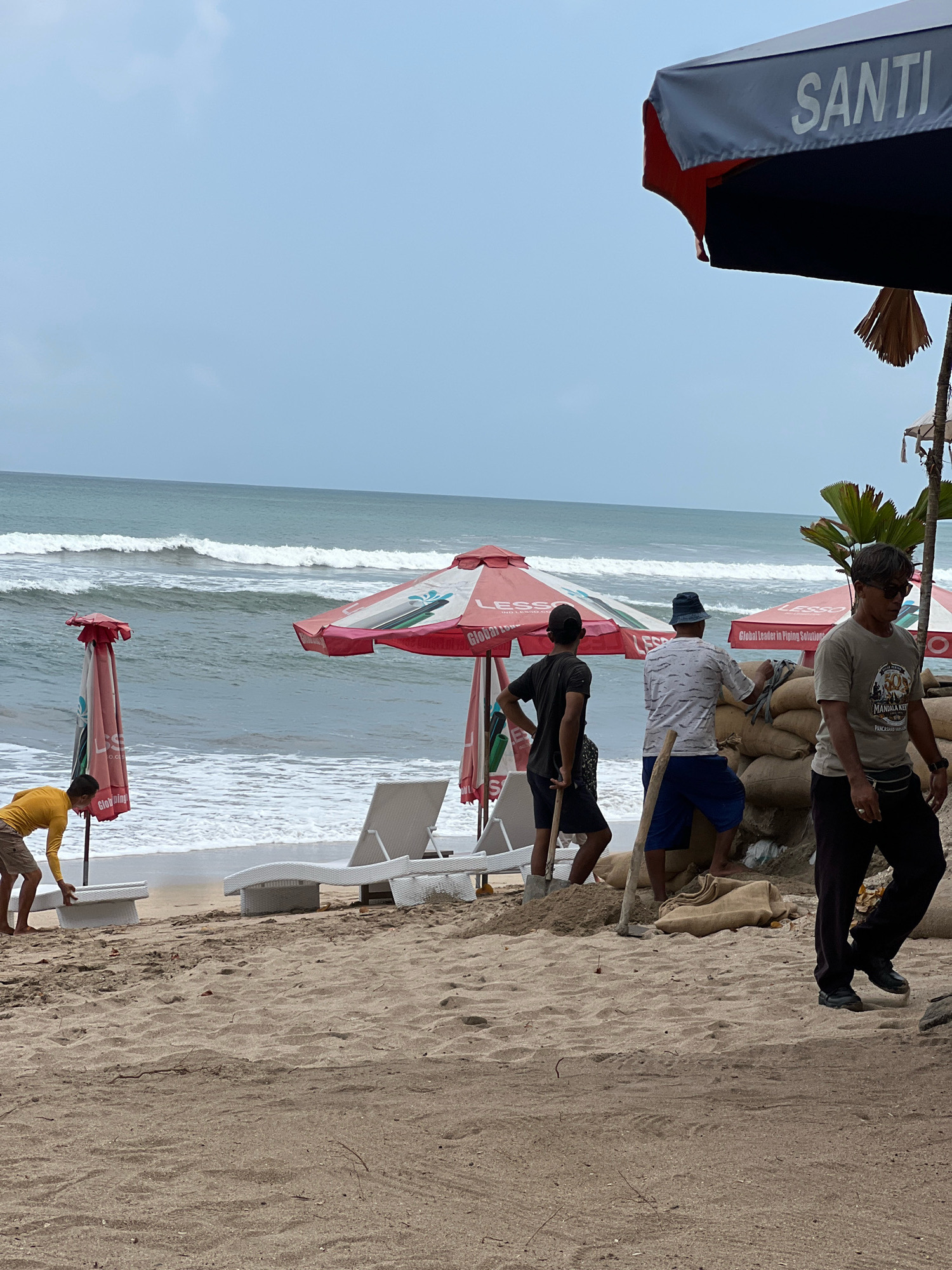 In Kuta, Indonesia, on a sandy beach, several people are working near the shoreline. Two men, one wearing a black t-shirt and the other a patterned white shirt and blue shorts, are actively shoveling sand into sandbags. A third man, in a grey t-shirt and dark pants, walks away from the sandbag fortification. In the background, the ocean waves are rolling in, and beach umbrellas and lounge chairs are visible. A person in a yellow shirt and shorts is seen arranging a white lounge chair near the water. The sky is overcast.