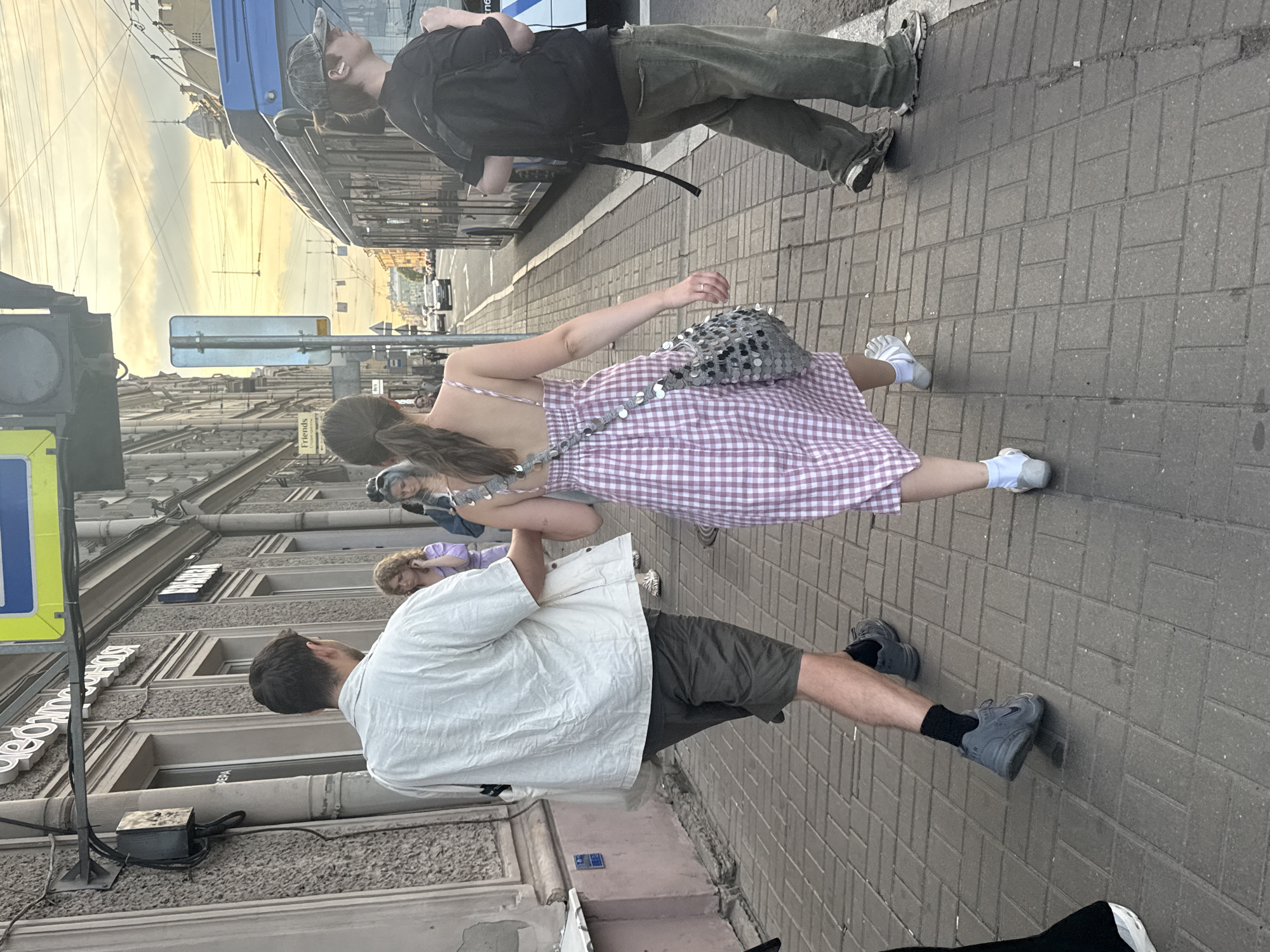 Two young adults, a man and a woman, walk along a pedestrian sidewalk in Saint Petersburg, Russia. The man, from behind, wears a light-colored short-sleeved shirt and dark shorts, with gray sneakers. The woman in front of him wears a pink and white checkered sundress with a sequined bag slung over her shoulder and white socks with light-colored shoes. They are walking away from the camera. To their right, a trolleybus travels down the street. The sidewalk is paved with light gray brickwork. Buildings line the left side of the street, with storefronts and windows visible. The sky above is a soft yellow and orange, suggesting it is late afternoon or early evening. A traffic light is visible in the upper left corner of the image. The overall scene depicts everyday urban life in Saint Petersburg.