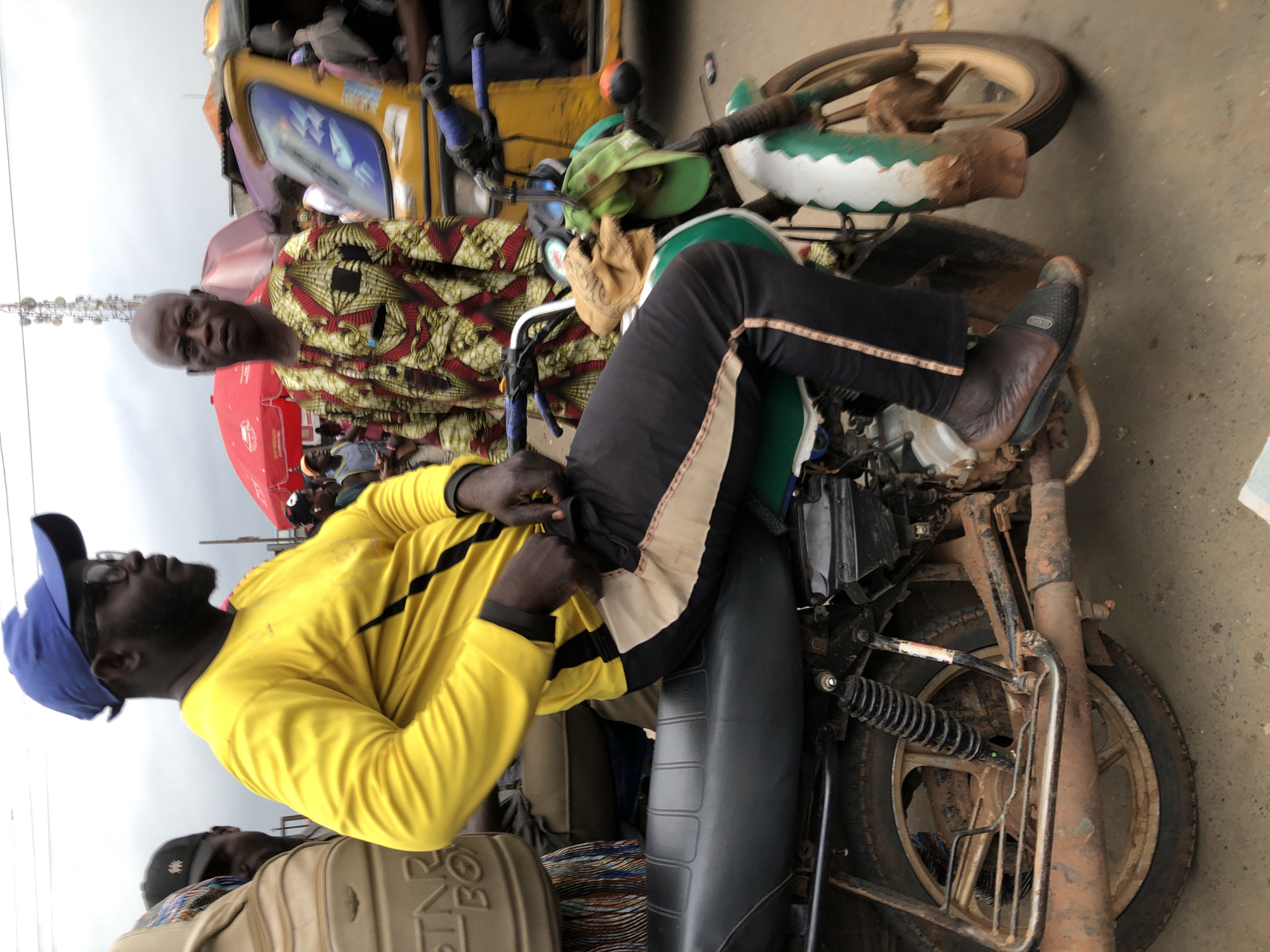 In Ikeja, Nigeria, on a cloudy day, a man wearing a bright yellow long-sleeved shirt and black pants with a beige stripe sits on a weathered motorcycle. He has a blue baseball cap and sunglasses on, and his hands are adjusting the fabric of his pants. Beside him, another man in a colorful, patterned robe looks towards the camera. The motorcycle, painted green and white, is covered in dirt. In the background, a yellow auto-rickshaw, red umbrellas, and a cell tower are visible, indicating a busy street market or transportation hub. The ground is a dirt road, suggesting a less developed area.