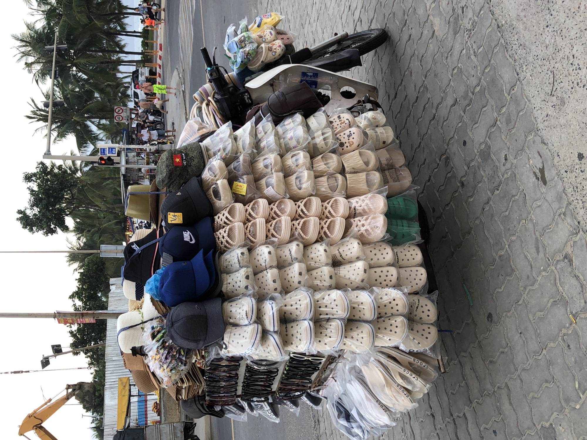 This image depicts a street scene in Đà Nẵng, Vietnam, focusing on a vendor's stall mounted on a scooter. The stall is heavily laden with an assortment of goods, primarily footwear and headwear. Rows of beige clogs, many packaged in plastic, are stacked vertically on the scooter's frame, along with various styles of sandals and flip-flops. On top of the footwear, an array of hats and caps are displayed, including baseball caps with brand logos like "NIKE" and "VF," as well as straw hats. Some of the hats feature Vietnamese flags or other decorative elements.
The scooter itself is parked on a paved street with a distinctive interlocking block pattern. In the background, a construction site with yellow heavy machinery and a white fence is visible. Further in the distance, palm trees line a sidewalk, suggesting a coastal or tropical setting. Several people are present in the background, including individuals on motorcycles and pedestrians, some appearing to be tourists. Traffic lights and street signs are also present, indicating an urban environment. The overall impression is of a vibrant street market operating in a busy city.
