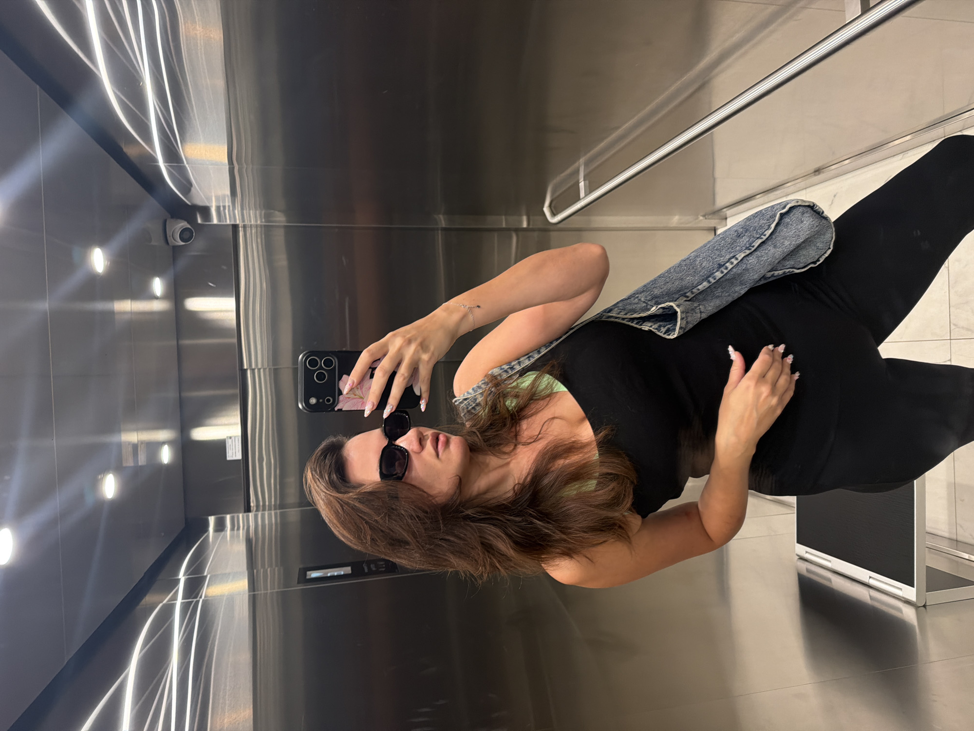 A woman is taking a selfie in an elevator. She is wearing a black tank top, black pants, and sunglasses. She has a denim bag slung over her shoulder. The elevator walls are made of stainless steel and there are lights on the ceiling. The floor of the elevator is tiled. A security camera is visible on the ceiling. The image is a typical selfie taken in an elevator, possibly in a modern building in Belgrade, Serbia.