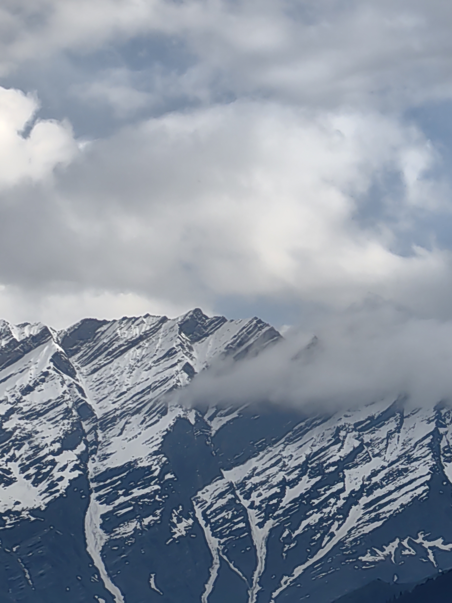 The image shows a close-up view of snow-capped mountains under a cloudy sky. The mountain slopes are steep and rugged, with visible striations of snow and dark rock. Some areas of the mountains are obscured by white clouds that appear to be low-lying, possibly obscuring the peaks. The sky is a mix of blue and grey clouds, with some brighter patches suggesting sunlight filtering through. The overall impression is of a high-altitude, rugged mountain landscape. There are no people or man-made objects visible in the image, and no specific details to indicate the location within Vashisht, India, beyond it being a mountainous region.