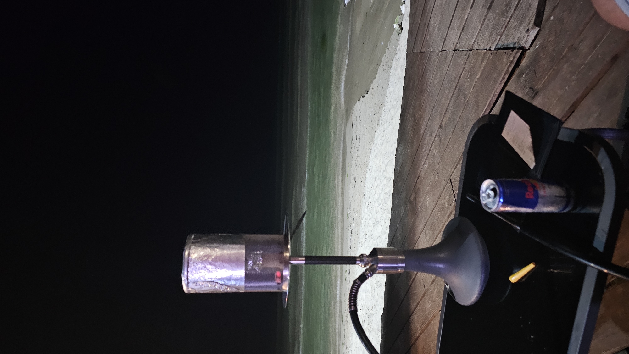 A hookah is set up on a table on a wooden deck overlooking a beach at night. The hookah base is dark gray, and the stem is silver and black. A black hose is attached to the hookah. The bowl of the hookah is covered with aluminum foil, and there appears to be hot charcoal on top. On the table, there is a Red Bull can, a yellow object, and a folded piece of paper. The beach is sandy, and the ocean is visible in the background with gentle waves. The sky is dark, indicating it is nighttime. The location is specified as the city of Varna, Bulgaria.