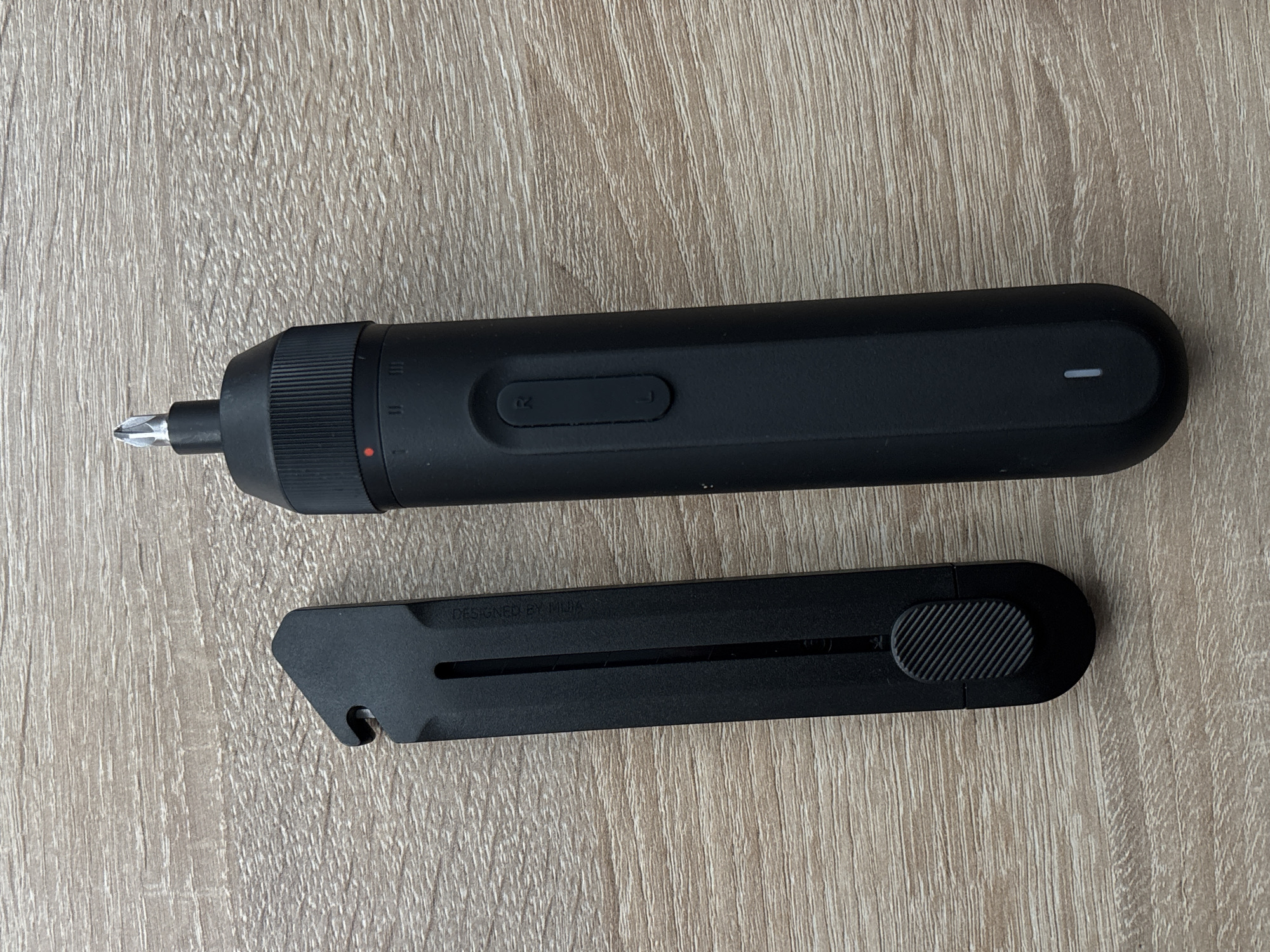 This image shows a top-down view of a black electric screwdriver and a black flat object, possibly a case or a tool, placed on a light brown wooden surface with a visible wood grain pattern. The screwdriver is elongated and cylindrical, with a silver Phillips head bit protruding from one end. It has a ridged grip section near the bit and a smooth body with two buttons and a small indicator light. The object below the screwdriver is rectangular and flat, with a cut-out slot in the center and a textured circular grip at one end. There is visible text on this object that reads "DESIGNED BY MUJA". The overall scene is simple and functional, with no people present. The lighting appears to be from above, casting subtle shadows. The context of Saint Petersburg, Russia, does not provide specific visual cues within the image itself, as the setting is generic. The image appears to be a product photograph or a casual snapshot of tools.