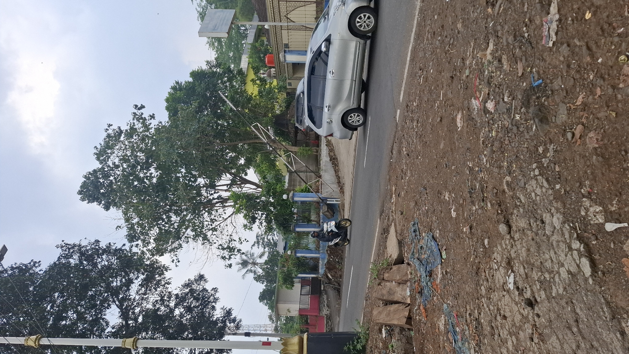 The image captures a street scene in Cilaku, Indonesia. A silver minivan is parked on the right side of the asphalt road, partially obscuring a large metal structure that appears to be a damaged street lamp or pole, leaning precariously over the sidewalk. A person is riding a white scooter on the road, moving from left to right, wearing a dark jacket. On the left side of the road, a white lamppost with golden decorative elements stands tall. Behind the lamppost and the leaning structure, there are buildings with red and pink walls and blue fences. The ground on the right side of the road is uneven and covered with dirt, rocks, and scattered debris, including blue netting and some plastic waste. The sky is overcast with clouds. The area appears to be a roadside with some vegetation and trees present.
