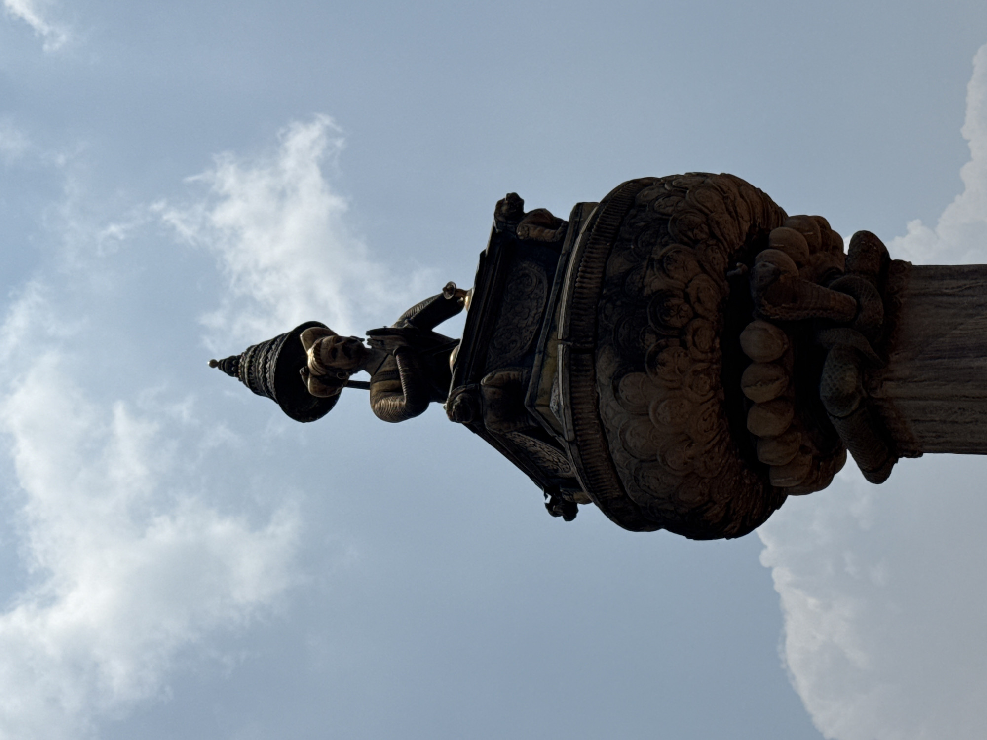 A low-angle shot depicts an ornate stone pillar against a cloudy sky. The pillar features intricate carvings, including a statue of a figure on top, adorned with what appears to be a pointed headdress and holding an object. The middle part of the pillar has elaborate designs resembling floral or geometric patterns, while the base appears cylindrical. The sky is a light blue with scattered white clouds. This pillar is likely located in Bhaktapur, Nepal, known for its rich cultural heritage and well-preserved ancient monuments.