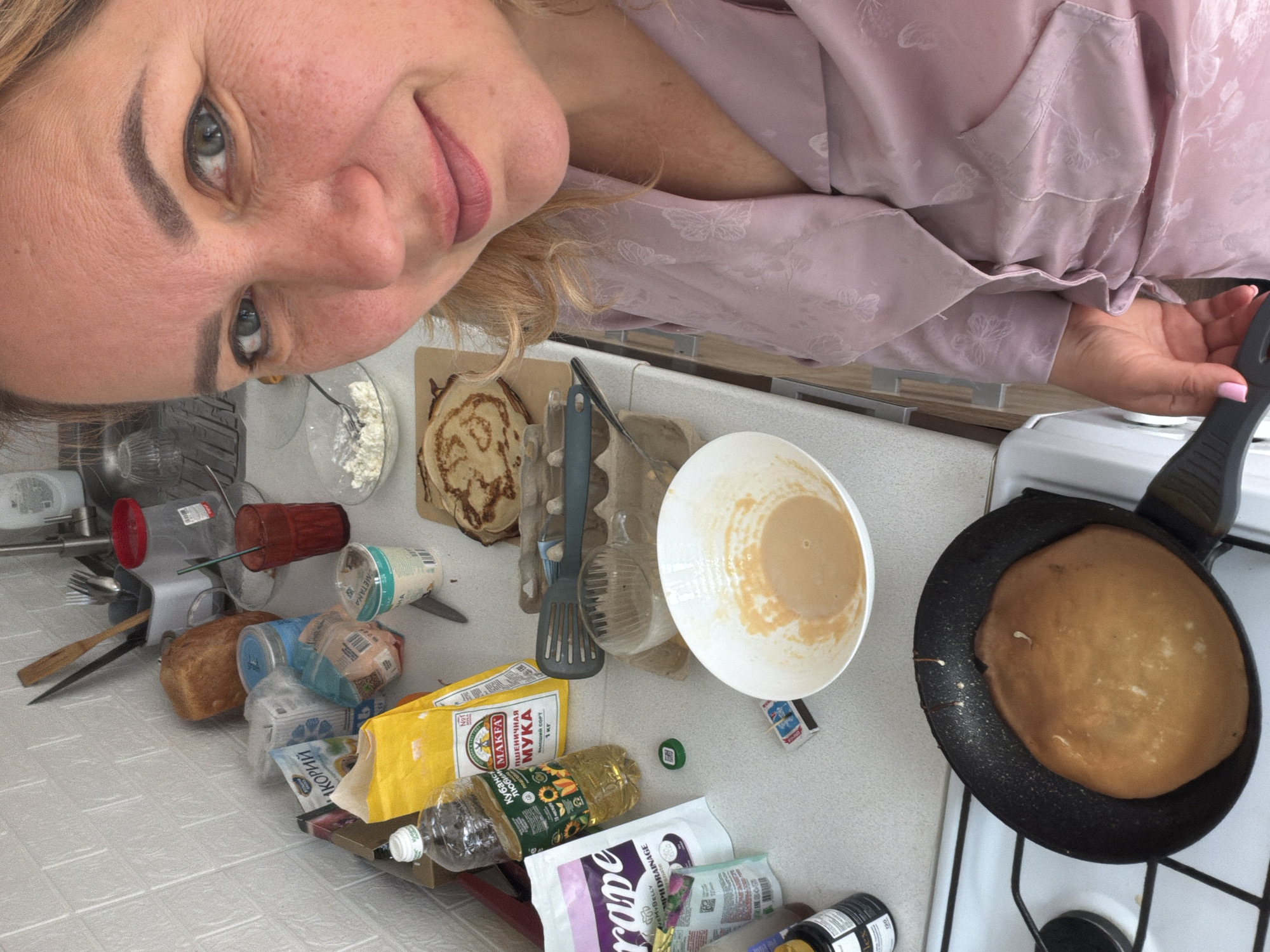 The image shows a woman looking at the camera with a slight smile, positioned over a cluttered kitchen counter. She is wearing a light purple, floral-patterned top. On the counter, there are various items: a frying pan with a pancake in it, a bowl with batter, cooking utensils, food packaging including a package of "MAKFA" flour, "Kuabnsk" oil, and other unidentified food containers. There is also bread, cutlery, and other miscellaneous items. Some finished pancakes can be seen on the countertop to the left. The scene appears to be a domestic kitchen setting. No evidence indicates this image was taken in the city of Leningradskaya, Russia. The scene lacks specific information that could determine its actual location.