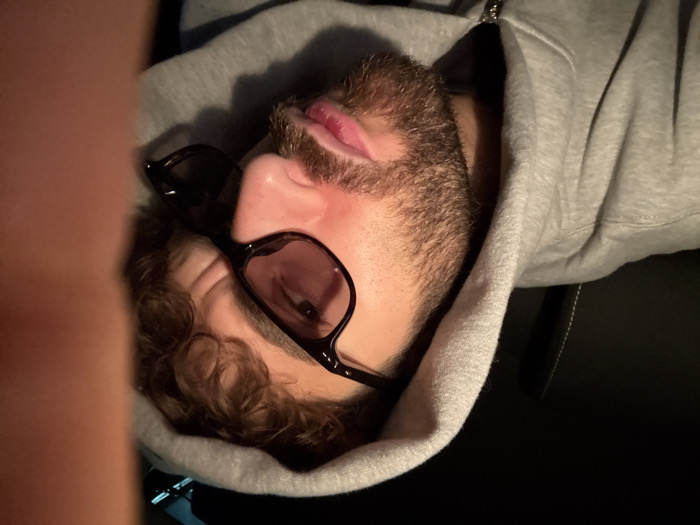 The scene depicts a close-up, angled portrait of a single adult male, reportedly located in Givors, France. The subject, positioned in what appears to be a dimly lit vehicle interior, occupies the majority of the frame. He has curly brown hair, a brown beard, and a mustache. He is wearing a grey hooded sweatshirt, the hood of which is pulled up and partially obscures his hair and head. Black-framed sunglasses with tinted lenses are worn, covering his eyes, and he appears to be looking directly or slightly downwards towards the camera. His mouth is slightly open, with his lips slightly parted. To the upper left, a dark vehicle seat with light-colored stitching is faintly visible. A blurred, flesh-toned object, consistent with a finger or thumb, obscures the rightmost portion of the image.