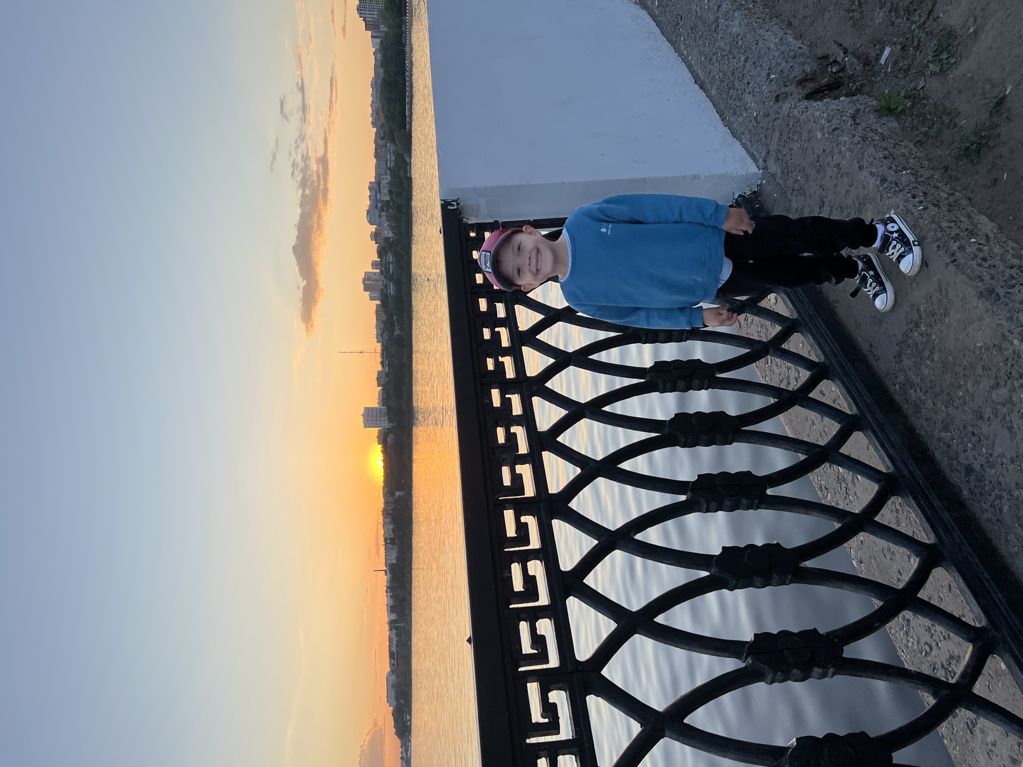 This image captures a young boy standing on an urban embankment in Izhevsk, Russia, during sunset. The boy, appearing to be between 4 and 7 years old, is positioned slightly right of center, facing the viewer with a broad smile. He is dressed in a blue long-sleeved shirt, black pants, and black sneakers adorned with a white star pattern on the sides and white soles. A red baseball cap is worn backward on his head.
To the boy's left, a dark, ornate metal fence featuring a Greek key-like pattern along its top edge and interconnected loops below runs along the edge of the concrete embankment. Behind the fence, a large body of water, likely the Izhevsk Pond or Reservoir, stretches across the midground. The water's surface exhibits gentle ripples and reflects the warm, orange, and yellow light of the setting sun.
On the distant bank of the water, a city skyline is visible against the horizon. The skyline comprises various buildings of different heights, along with a tall, slender antenna-like structure on the far left. The sun is prominently visible as a bright orange-yellow disc low on the horizon, slightly to the right of the image's center, casting a strong glow. The sky transitions from vibrant orange and yellow near the horizon to a pale blue overhead, with scattered, lightly colored clouds present, particularly in the upper quadrants. To the boy's immediate right, a plain white wall extends along the embankment. The ground beneath the boy is a dark, rough concrete surface.
