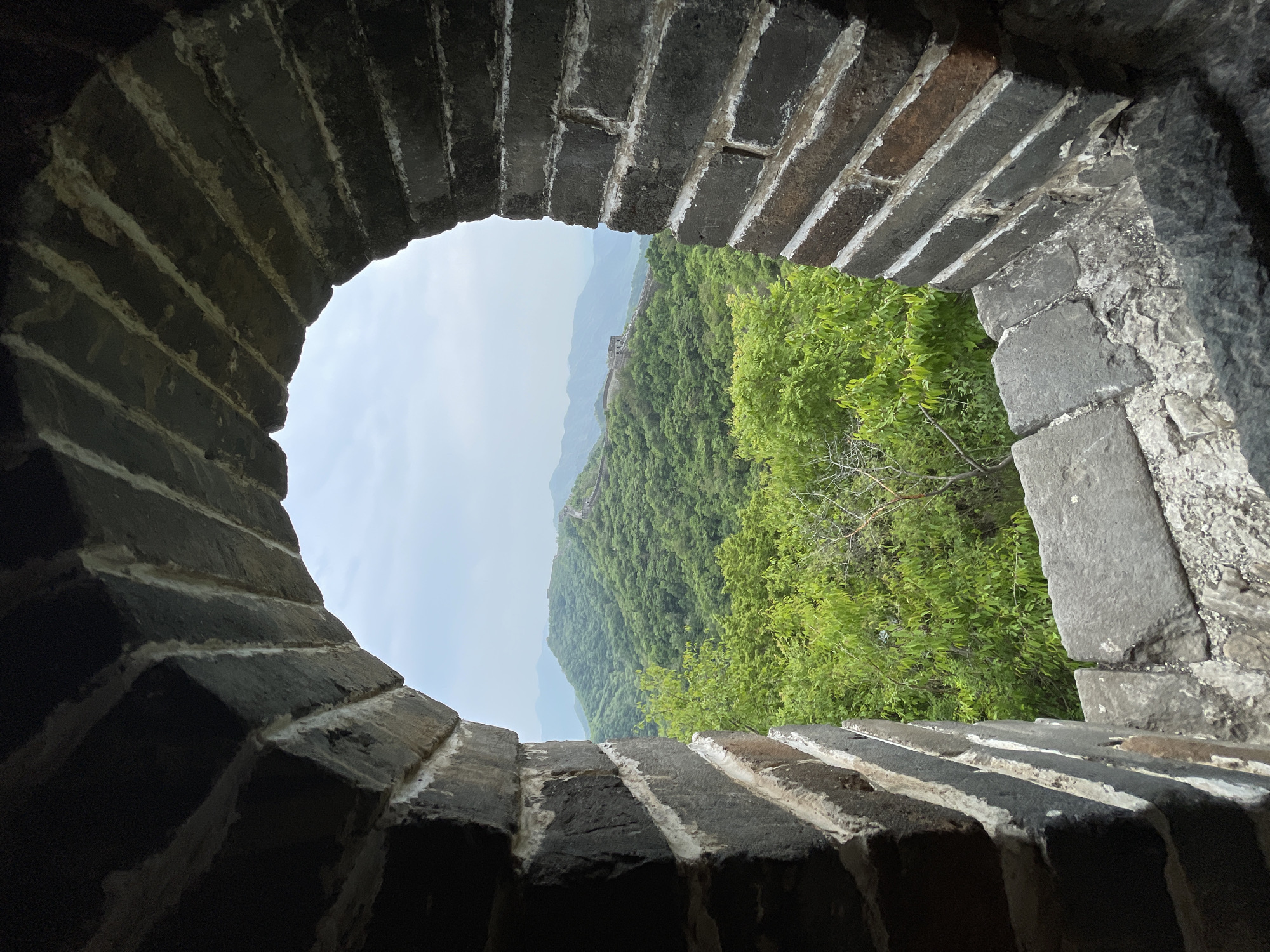 The image shows a view of a lush, green mountain range through a circular opening in a stone structure. The opening is made of dark grey bricks with white mortar. In the distance, the faint outline of what appears to be a wall or structure can be seen winding along the ridgeline of the mountains. The sky is overcast. The image was taken in the city of Huairou District, China.