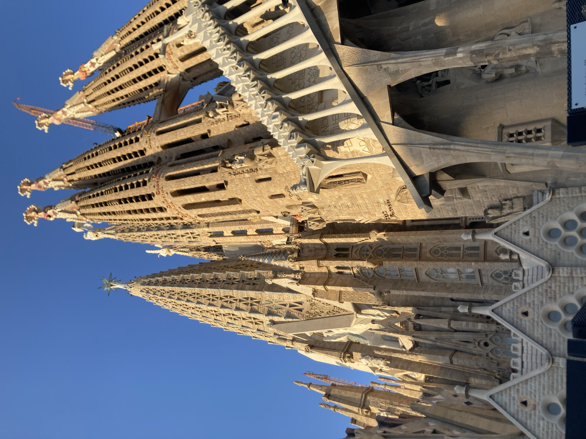 The image shows a low-angle, full shot of the Sagrada Familia in Barcelona, Spain, under a clear blue sky. The iconic basilica's intricate, unfinished spires and facades dominate the frame, bathed in warm, late afternoon sunlight. Cranes are visible among the upper spires, indicating ongoing construction. The foreground features detailed stone carvings and architectural elements, including arched structures and triangular rooflines. The building's stone is a warm, golden-tan color. No people are visible in the image.