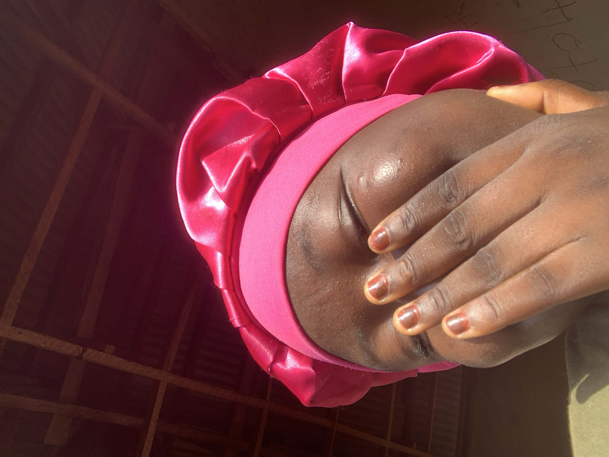 A close-up shot shows a young Black individual wearing a bright pink satin bonnet. Their eyes are closed, and their face is partially obscured by their hand, which is placed over their mouth. The individual's nails are painted a reddish-brown color. The background consists of a dimly lit interior with a wooden ceiling structure visible, suggestive of a simple dwelling. Sunlight streams in from the right, illuminating the side of the individual's face and creating highlights on their skin. The scene appears to be in Jalingo, Nigeria.
