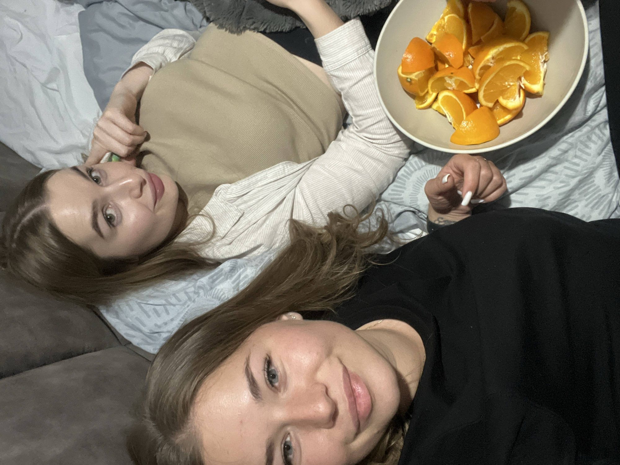 Two young adult females are shown indoors, reclining on what appears to be a bed or sofa. The individual in the foreground, positioned on the left, has light brown hair, blue eyes, and is wearing a black long-sleeved shirt. She is looking directly at the camera with a slight smile. Her right hand, featuring white-painted fingernails, is visible near a white ceramic bowl. This bowl contains multiple cut segments and wedges of oranges. The second individual, positioned behind the first and slightly to the right, has brown hair, green/hazel eyes, and is wearing a light-colored, vertically striped button-up shirt over a tan ribbed top. She is also looking towards the camera with a slight smile, with her left hand raised towards her face, displaying a green-painted index fingernail. The background includes a dark gray upholstered surface on the left, light blue patterned bedding, and a dark gray fluffy blanket on the right. The specific location in Budva, Montenegro, cannot be determined from the visual evidence.