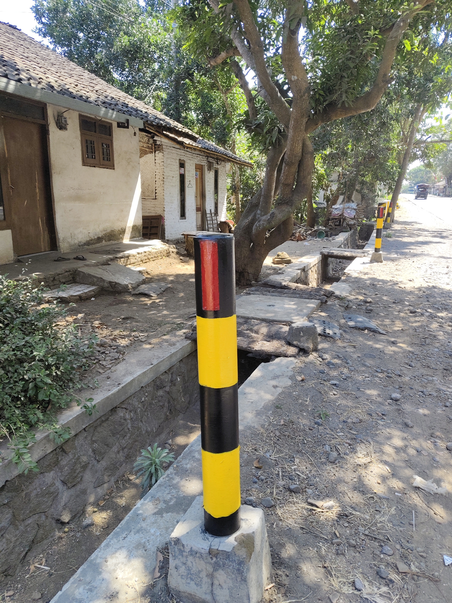 A roadside scene in Gambirono, Indonesia, depicts a weathered building and a tree-lined street under a bright sky. In the foreground, a black and yellow striped bollard stands on a concrete base, with a stone-lined ditch beside it. The building, appearing to be a house or small shop, has a tiled roof, white plastered walls, and wooden doors and windows. Adjacent to the building, lush green foliage grows. Further down the street, more yellow and black bollards mark the roadside, and a few vehicles are visible in the distance. The ground is a mix of dirt, gravel, and scattered debris. The overall impression is one of a rural or semi-urban environment with signs of age and natural elements.