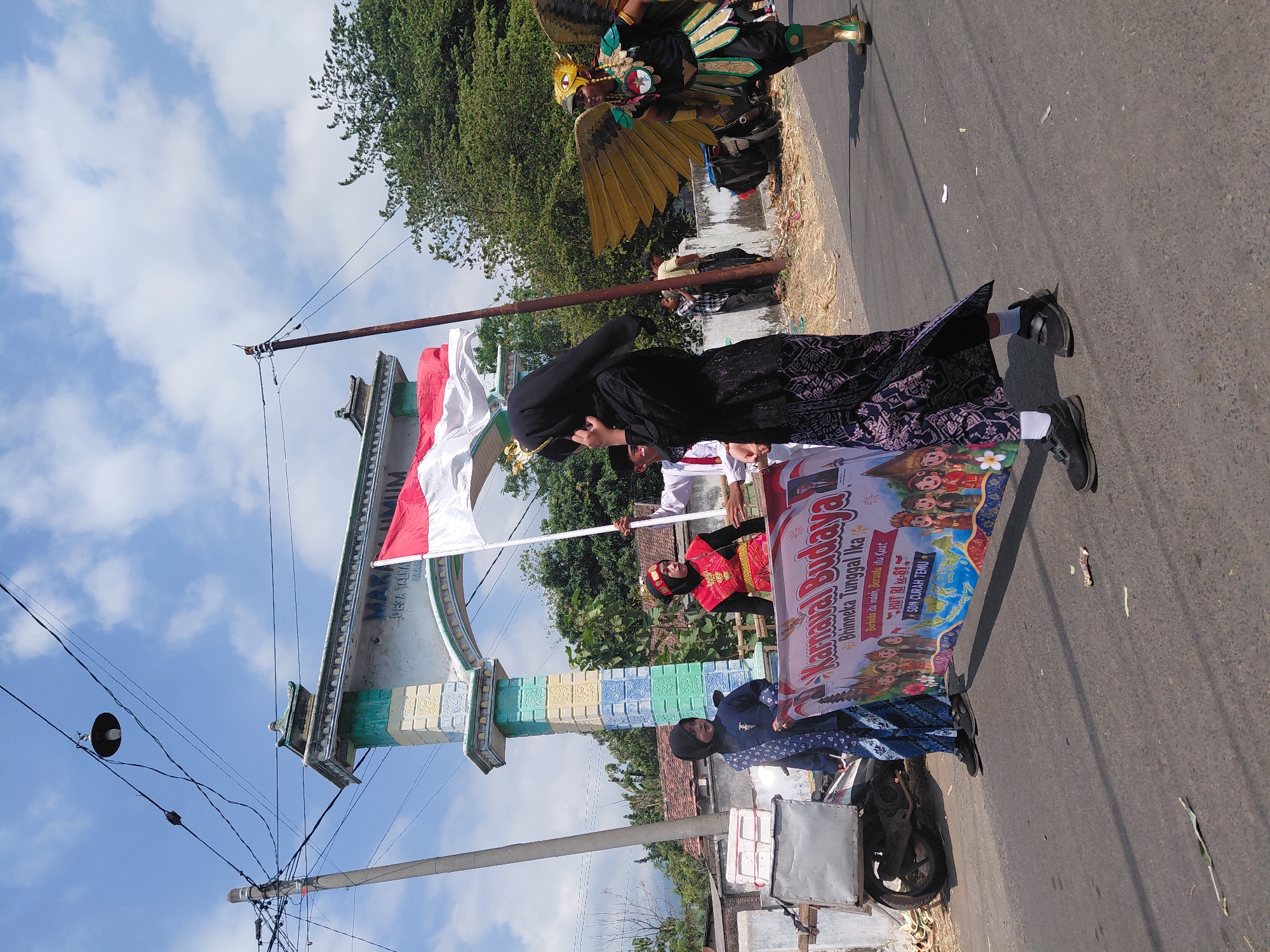 In the city of Bimo, Indonesia, on a sunny day with a blue sky and scattered clouds, a cultural parade is underway. A group of participants is walking down a paved road, some carrying a banner that reads "Karnaval Budaya" (Cultural Carnival) and "Bhinneka Tunggal Ika" (Unity in Diversity). One person in the foreground is wearing a black hijab and a patterned skirt, holding the banner. To their left, two children are dressed in traditional blue and white attire. To the right, a person in a colorful costume with large golden wings, resembling a mythical bird, is marching. In the background, an archway with intricate designs marks an entrance, and trees line the side of the road. Power lines and a street lamp are visible above the scene. Several motorcycles are parked on the side, and a handcart is also present. The overall atmosphere suggests a festive community event.