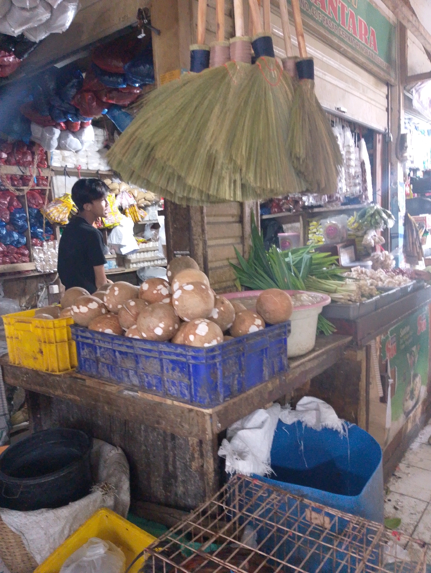 This is a medium shot of a street market scene in Lembang, Indonesia. In the foreground, a young man wearing a black t-shirt is standing and looking to the right. He is partially obscured by a large bunch of brooms with straw bristles and wooden handles, which hang from a wooden structure above. In front of the man, a yellow plastic crate sits on a wooden table. To the right of the yellow crate, a blue plastic crate is filled with coconuts. Behind the coconuts, there are some green onions and other vegetables laid out on a dark surface, and further to the right, a green sign with red lettering is visible. In the background, shelves are stacked with various goods, including plastic bags filled with items, and more brooms are hanging. The overall environment appears to be an outdoor or semi-outdoor market stall.