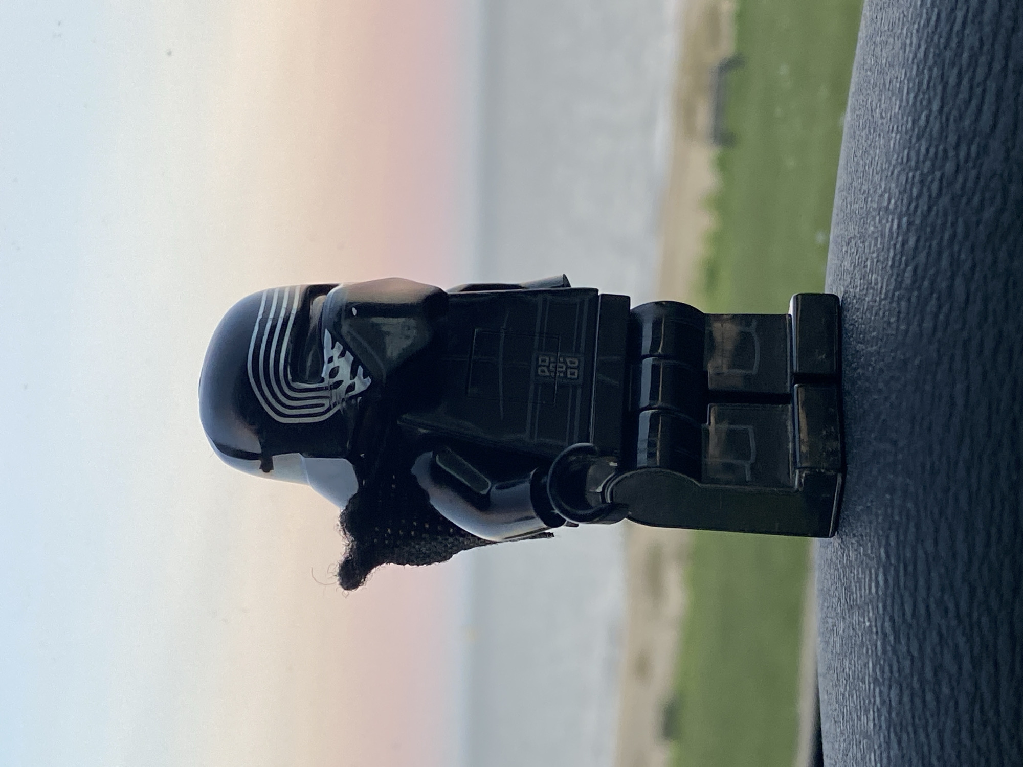 A close-up, eye-level, vertical shot depicts a black Lego Kylo Ren figure resting on a dark, textured surface, likely a car dashboard, with a blurred background showing a horizon of water and green land under a light-colored sky. The figure is positioned horizontally in the frame with its helmet and upper body on the left and its legs on the right. The helmet's details and markings are visible. The figure appears stationary. Due to the lack of identifiable landmarks in the background, it's not possible to confirm the specific location within East Suffolk, United Kingdom, or even be certain of the country.