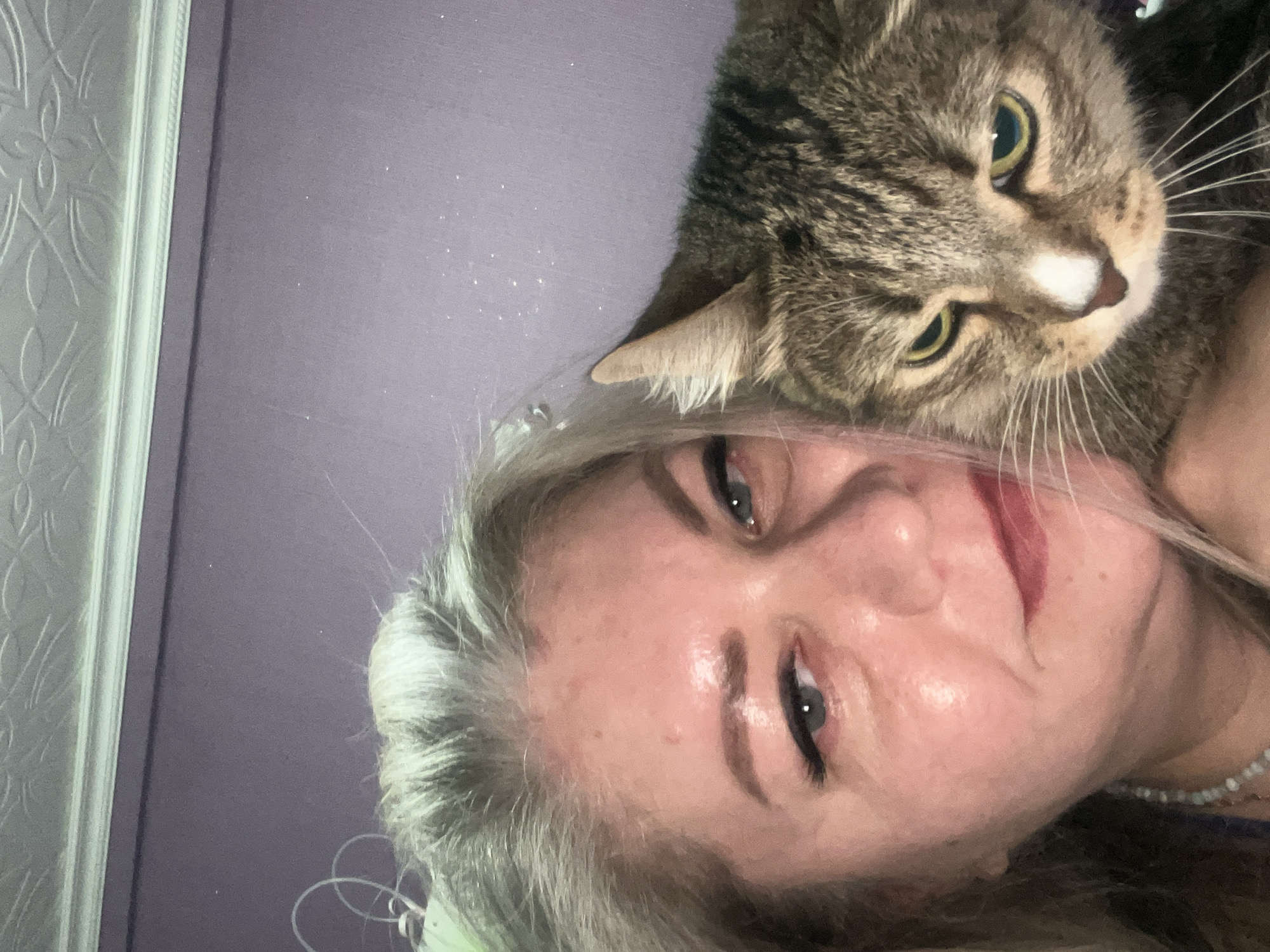 A woman with fair skin and gray hair is holding a brown tabby cat with green eyes. The woman is looking at the camera and has red lipstick. The cat is looking slightly to the right. The background is a light purple wall. A door frame is visible on the left side. The location is not identifiable in the city of Izhevsk, Russia, without more context.