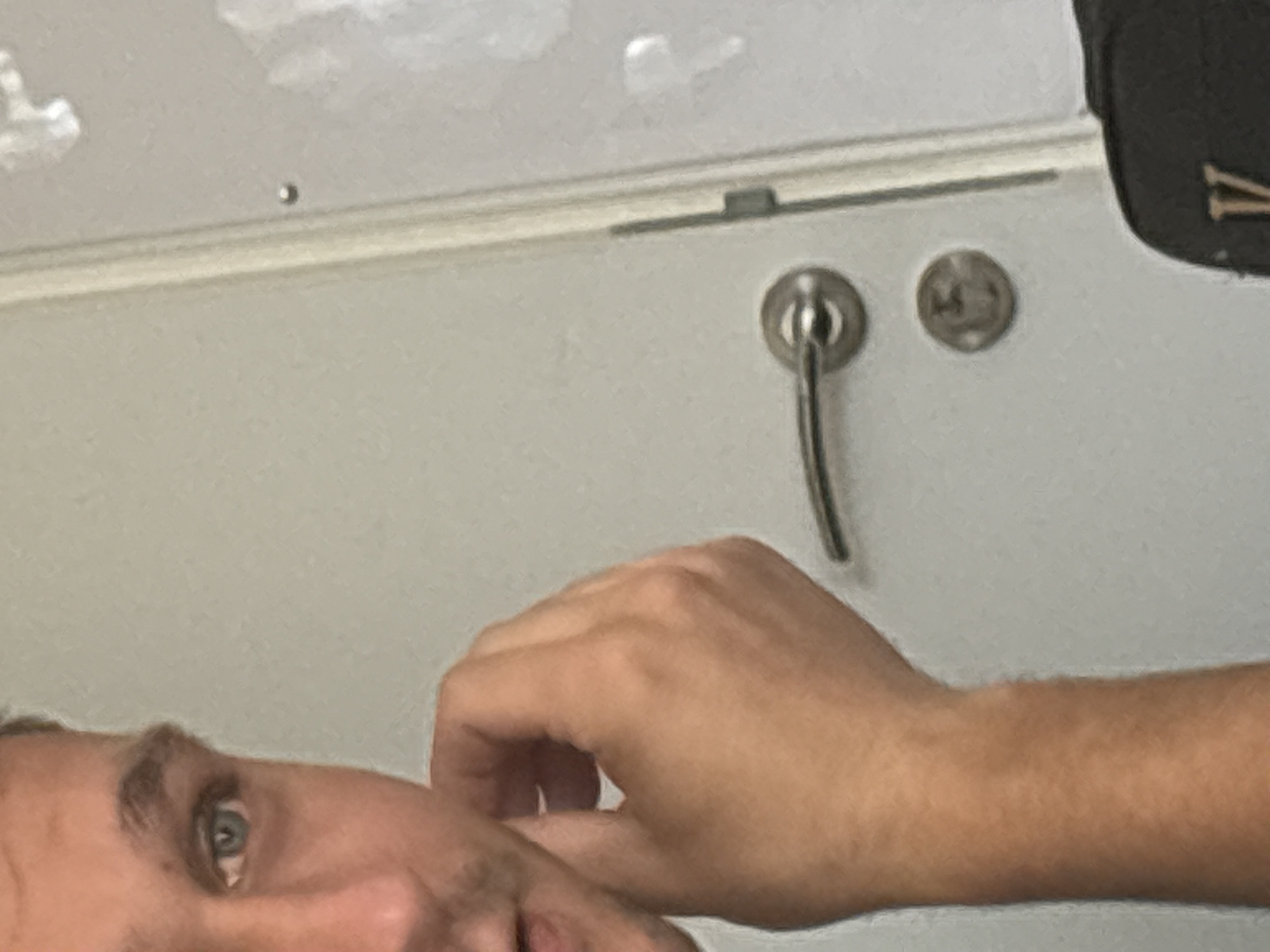 The image is a close-up, waist-up shot of a person's face and arm. The person, whose gender is not discernible, has light skin and blue eyes. Their left hand is raised to their face, touching their cheek. The background is a plain, light-colored wall with a silver door handle and a small silver disc. The lighting is even, and the image is slightly out of focus, suggesting it was taken with a smartphone camera. The image is likely a selfie, given the framing. There are no discernible landmarks or contextual clues to indicate a specific location within a city.