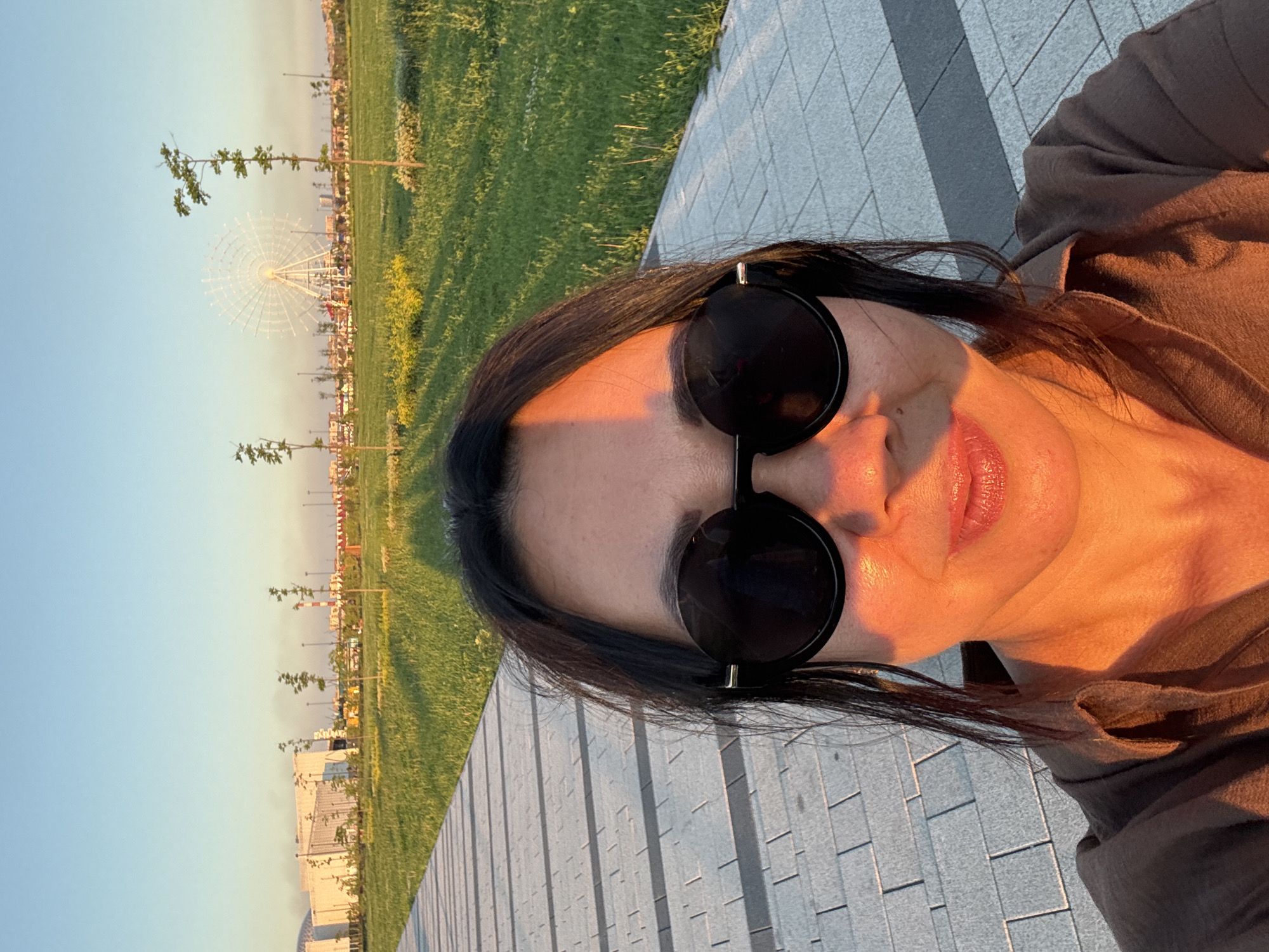 A woman with dark hair and sunglasses is taking a selfie in an outdoor park area. She is wearing a brown shirt. The background shows a paved walkway, a grassy field with scattered trees and yellow flowers, a Ferris wheel, and some buildings. The lighting suggests it is late afternoon or early evening, with warm sunlight casting shadows. The scene appears to be a modern park or recreational area within a city. The city of Magnitogorsk, Russia, is known for its industrial history and has been undertaking urban renewal projects, which could include areas like the one depicted.