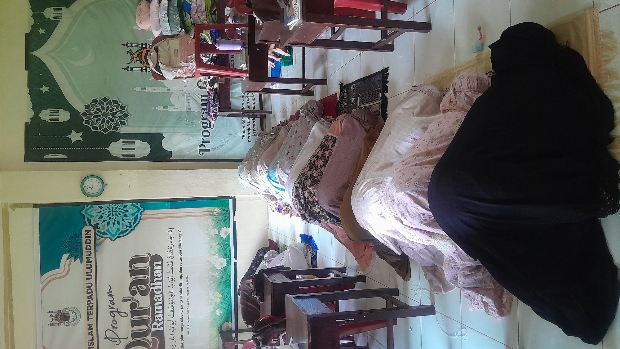 A group of Muslim women is praying in a room in Lhokseumawe, Indonesia. They are kneeling on prayer mats, facing towards a wall. The woman in the front is wearing a black niqab. Other women are wearing pink and white colored abayas. Behind them are small brown wooden desks with red chairs. On the wall behind the women, there is a green and white banner with a mosque design and text in Arabic and Indonesian related to Ramadan. There is also a green and white program banner with the words "Program Quran Ramadhan." A clock is on the wall above it.