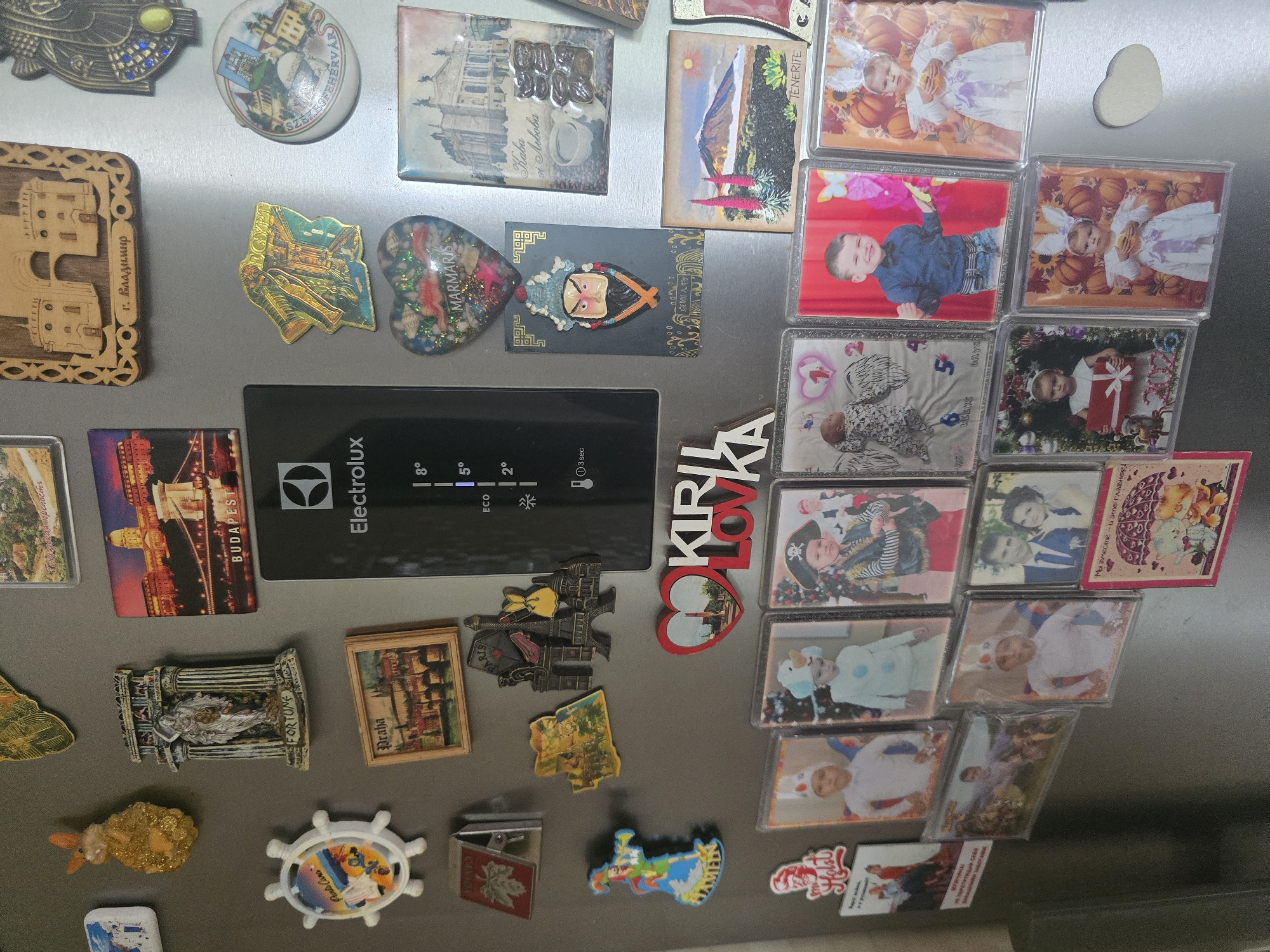 The image shows a close-up of a refrigerator door covered with various magnets and photographs. The refrigerator appears to be in a domestic setting, as suggested by the collection of personal items.
Prominent magnets include:
- A large black Electrolux appliance panel displaying temperature readings (8°, 5°, 2°) and an "Eco" setting.
- A yellow glittery rabbit magnet.
- A rectangular magnet depicting a building labeled "BUDAPEST".
- A metallic souvenir magnet of the Eiffel Tower and a street scene.
- A heart-shaped magnet with "MARMARIS" written on it, containing shells and glitter.
- A magnet with a Chinese opera mask.
- A magnet with a depiction of a mountain and flowers labeled "TENERIFE".
- A circular magnet with a depiction of a town and a church.
- A magnet of a classical building.
- A magnet of a clown with a trumpet.
- A magnet with a vineyard scene.
- A magnet shaped like Egypt.
- A magnet shaped like a building with a keyhole.
- A magnet with a picture of a person playing an instrument in a field.
Numerous photographs, mostly of children, are also attached to the refrigerator door. These include:
- Several photos of children in costumes, such as a pirate, a snowman, and holiday-themed outfits with Christmas trees and pumpkins.
- A photo of a baby with angel wings.
- A wedding photo of a couple.
- A photo of a young boy in a blue shirt.
- A photo of a young girl in a white dress with bunny ears.
Other items include:
- A red magnet with a white leaf design.
- A heart-shaped cutout magnet.
- A pink magnet with text that appears to be in Cyrillic script, featuring cartoon bears and an umbrella.
- A red magnet with the word "Holst" in a stylized font.
- A magnet with a picture of people on a bus.
- A small wooden magnet with a house shape.
The overall arrangement suggests a collection of travel souvenirs and personal mementos. The lighting is bright, indicating the photograph was taken with artificial light or in a well-lit environment.