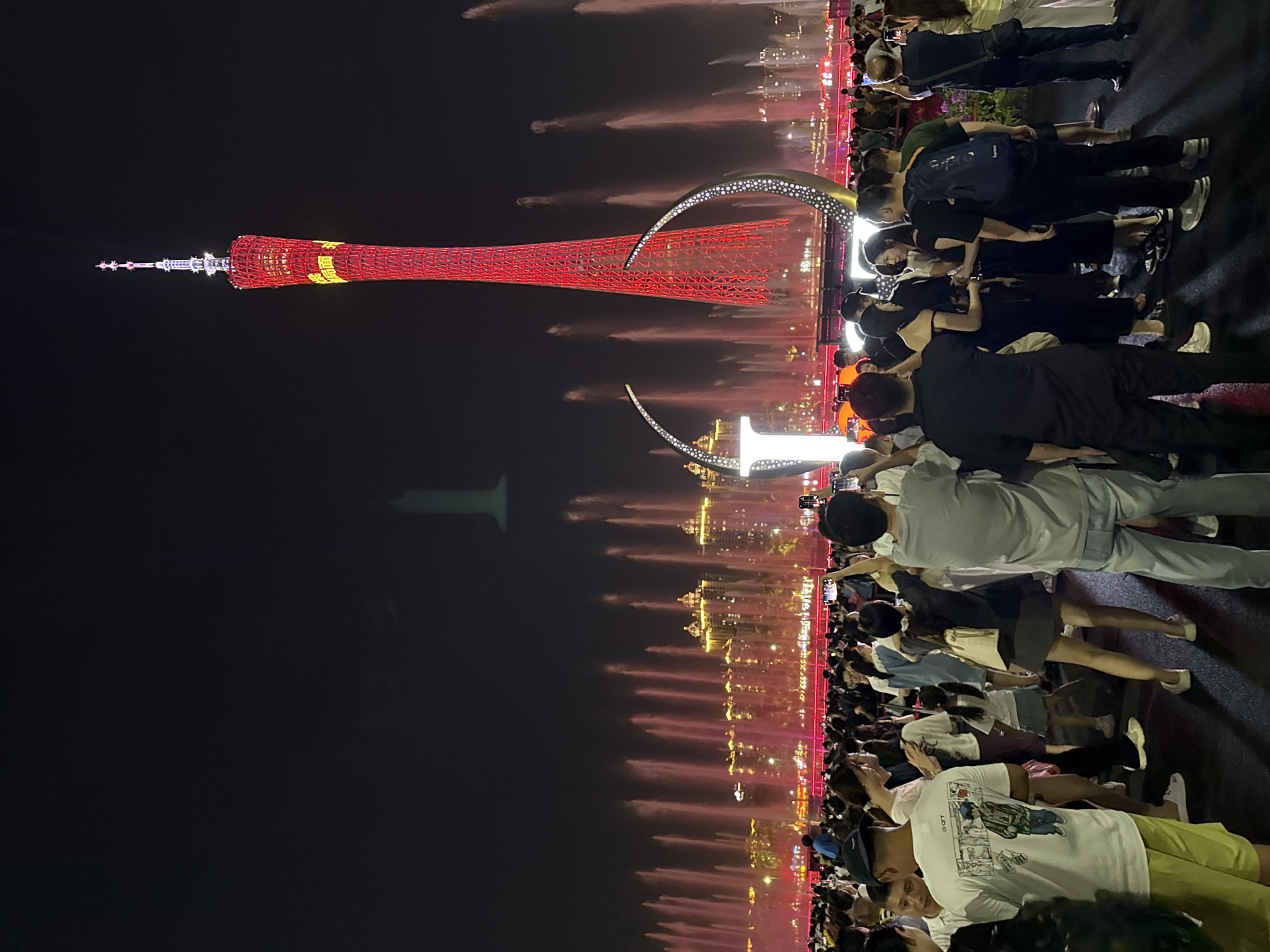 In Tianhe District, China, a night scene unfolds with a prominent red, lattice-like tower illuminated and dominating the sky. Surrounding the tower, a large crowd of people gathers, many holding up their phones to capture the spectacle. In the background, a synchronized fountain show erupts with numerous water jets, back-lit in pinkish-red, creating a vibrant display. Buildings with illuminated windows are visible behind the fountains, and large, abstract, metallic sculptures with dotted patterns are positioned in the foreground, adding to the festive atmosphere. The ground is paved, and the overall impression is one of a public gathering enjoying a nighttime event featuring light displays and water features.