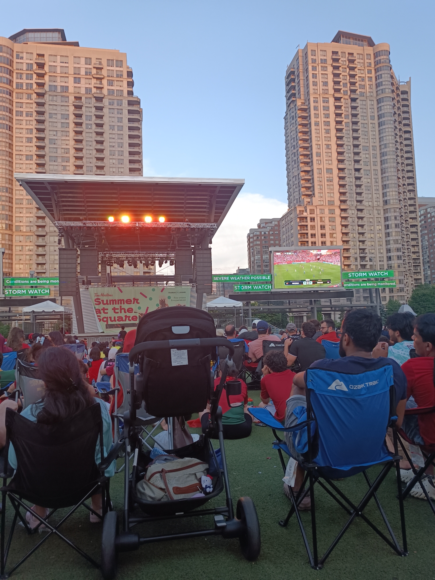 This image depicts an outdoor event in Celebration Square, Mississauga, Canada. A large crowd is gathered, seated in portable chairs on a green turf surface, facing a stage and a large video screen. The stage is set up with lighting equipment. The video screen displays a soccer game, with team names "PORTUGAL" and "GHANA" visible. Several tall residential buildings form the backdrop of the scene. Two prominent "STORM WATCH" signs are visible, one on each side of the stage, indicating a weather monitoring system in place. A stroller is positioned in the foreground, adding a family-oriented element to the gathering. The overall atmosphere suggests a public event, possibly a community screening of a sports match or an outdoor concert.