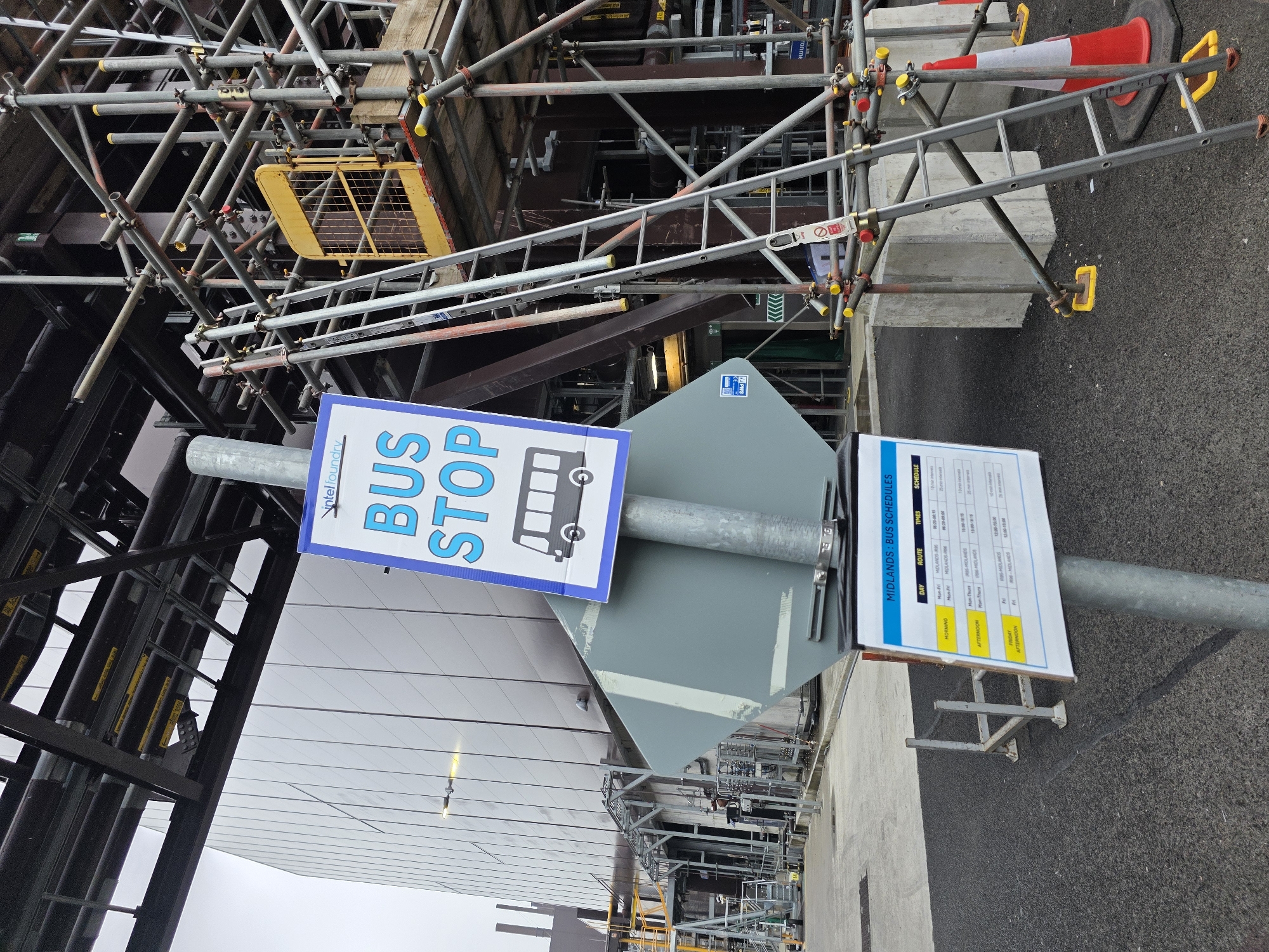 The image depicts a construction site in Leixlip, Ireland, adjacent to an Intel facility. In the foreground, a metal pole supports a "BUS STOP" sign with a graphic of a bus. Below this, a laminated schedule titled "MIDLANDS: BUS SCHEDULES" details bus routes, times, and intervals for weekdays and Friday afternoons. The background is dominated by the modern, grey-paneled exterior of the Intel building. To the right, extensive scaffolding covers another section of the industrial complex, with a ladder leaning against it. A bright orange traffic cone is also visible near the scaffolding. The ground is paved with asphalt, and the overall atmosphere suggests an active industrial or construction environment under an overcast sky. No people are present in the frame.