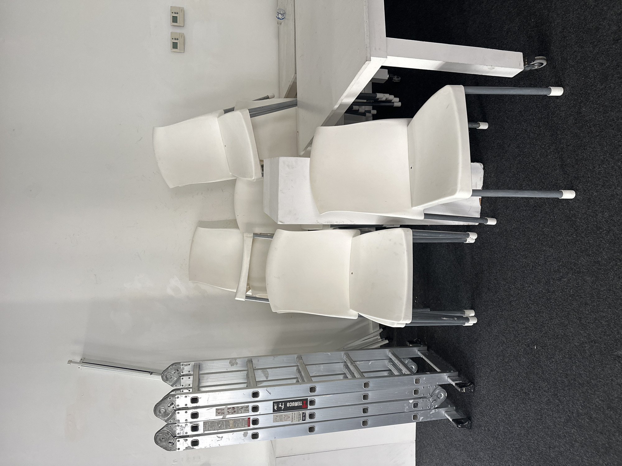 The image depicts a section of a room with white walls, grey carpet and white furniture. Stacked against the wall are three white plastic chairs with grey metal frames. A closed aluminum extension ladder is leaning against the wall near the chairs. The chairs are stacked on top of each other and are positioned next to a white table. There are small electrical outlets on the wall. Based on the style of electrical outlets, it is possible, but not confirmable that this photo was taken in Buenos Aires, Argentina.
