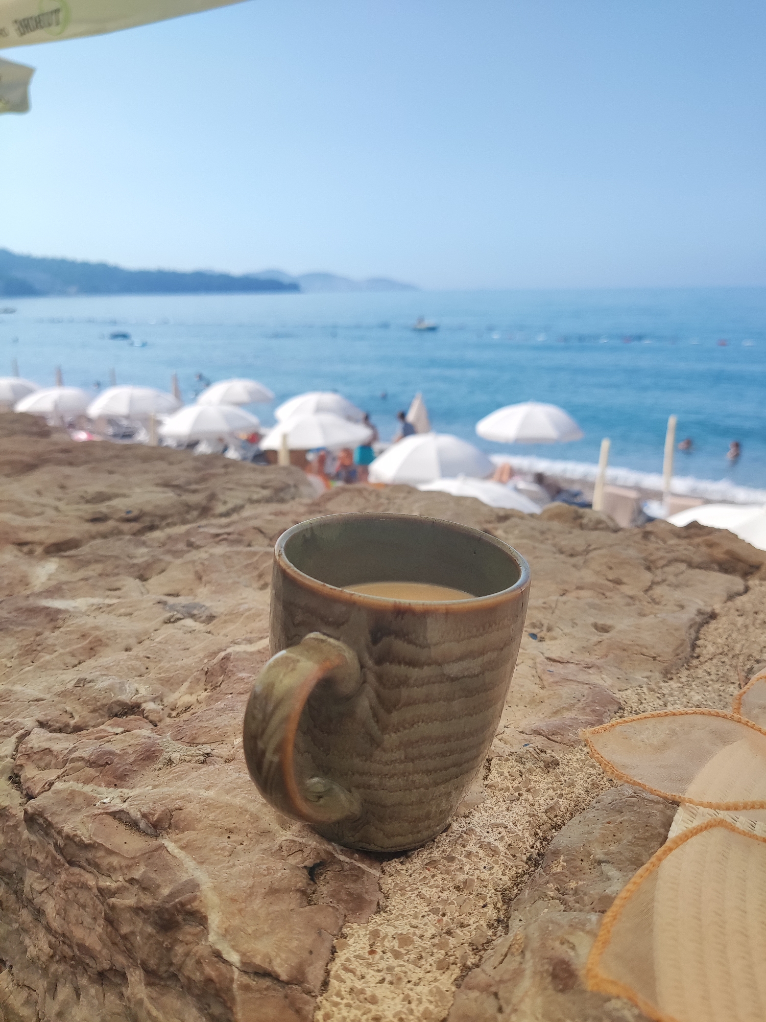 A close-up, eye-level shot of a ceramic mug filled with a light brown liquid sits on a textured, rocky surface. The mug, a muted green with horizontal striations and a curved handle, is positioned slightly off-center to the right. The rocky surface appears to be a natural rock formation, with variations in color and texture. In the background, out of focus, is a beach scene. The ocean is a clear blue, meeting a sandy shore dotted with numerous white umbrellas. Several people are visible in the water and on the beach, suggesting a public recreation area. The sky is a clear, pale blue. The overall impression is a relaxed, seaside setting, likely during the daytime. The context implies this is a scene from Sutomore, Montenegro, a popular coastal town known for its beaches.