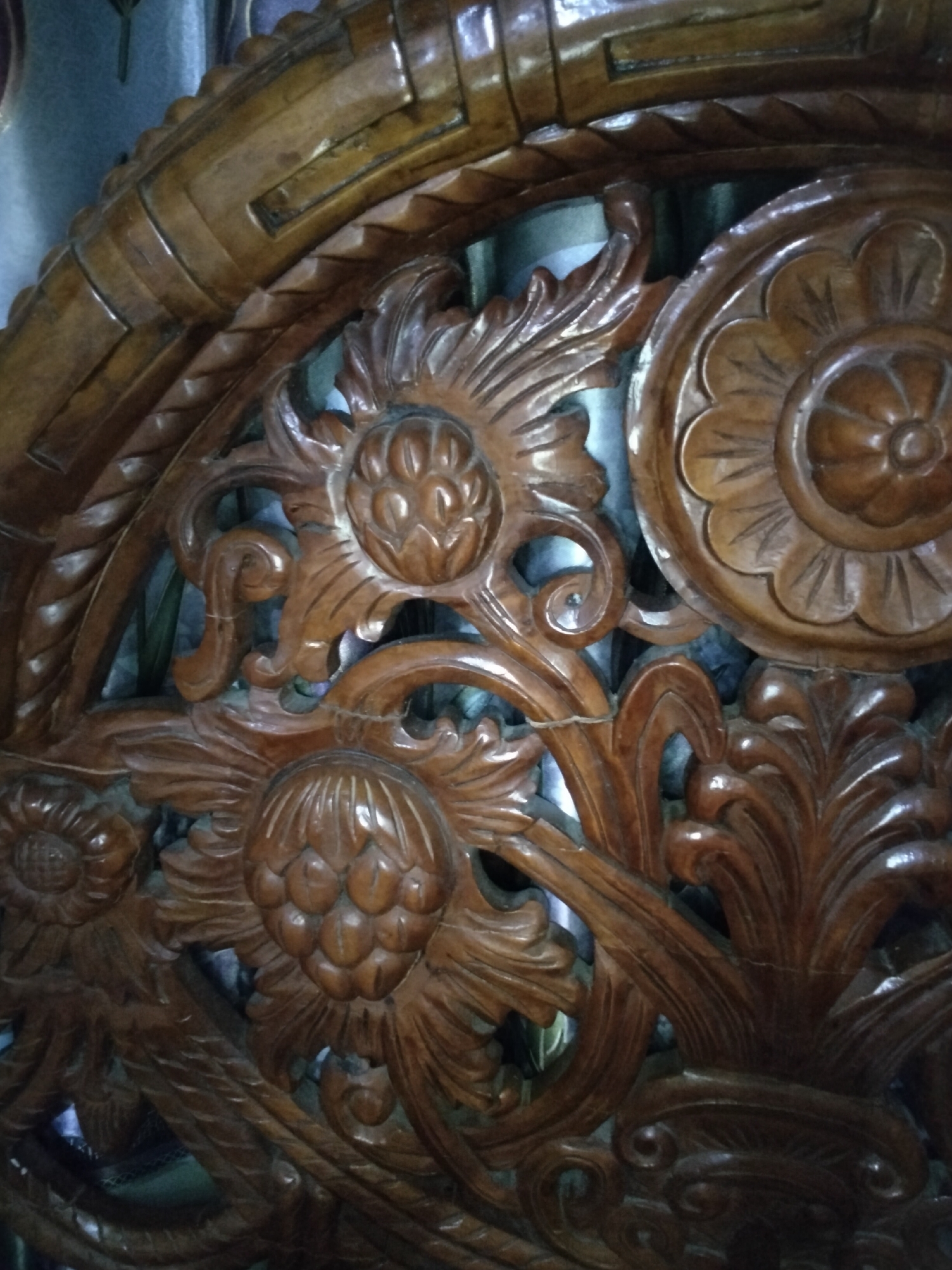 The image is a close-up, overhead shot of intricately carved dark wood. The carving features floral motifs and swirling leaf patterns, rendered in a relief style. The wood appears to be polished and has a warm, reddish-brown hue. The texture of the wood is evident, with smooth, curved surfaces and sharp, defined edges in the carved details. Some areas of the carving show a slightly lighter shade, suggesting wear or variations in the wood grain. In the background, a dark green curtain is visible through some of the carved openings. The lighting casts subtle shadows, highlighting the depth and dimension of the carvings. This object is located in Payagaboh, Indonesia.