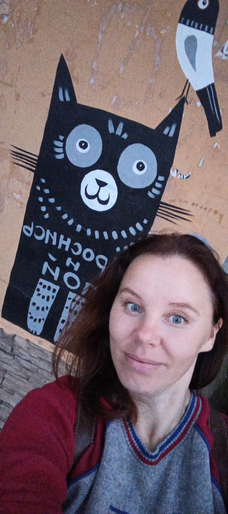 In Votkinsk, Russia, a woman with auburn hair and blue eyes takes a selfie in front of a mural. The mural features a stylized black cat with large gray eyes and a white mouth, positioned on a tan, textured wall. Above the cat, to the right, is a black and white bird. The cat's body is decorated with white polka dots and a pattern resembling stitching. Text, appearing to be in Cyrillic letters and oriented upside down from the viewer's perspective, is visible on the cat's body. The woman is wearing a red hooded sweatshirt over a grey sweater with red and blue trim around the V-neck. She is smiling, with her head tilted slightly. The background wall shows signs of wear and tear.