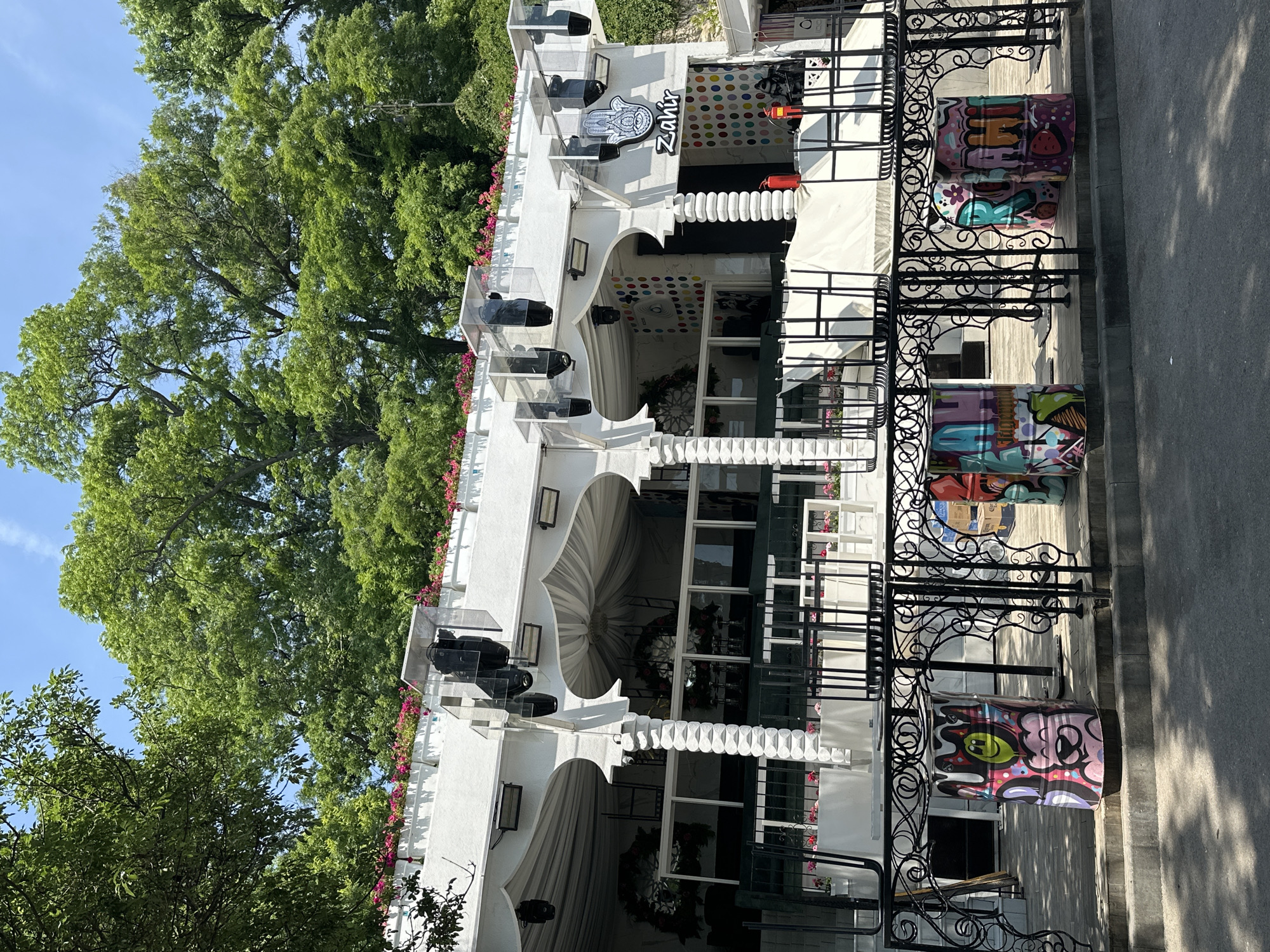 A bright daylight exterior shot captures the facade of a white, multi-level building with an ornate black metal railing in the foreground. The building exhibits an architectural style featuring multiple arches and decorative columns with a stacked disc pattern. The upper perimeter of the building displays white planters filled with vibrant pink flowers. Above the arches, several black stage lights or speakers are mounted under clear protective enclosures.
On the right side of the building, a prominent white sign depicts a Hamsa hand symbol above the word "Zahir" in black stylized text. Below this, and extending to an adjacent section, the wall is adorned with a colorful polka-dot pattern. Red fire extinguishers are visible at the base of this wall. Through the arches, draped white fabric ceilings and some decorative elements, including what appears to be a floral wreath, are discernible. White outdoor furniture, possibly stacked tables and chairs, is visible behind the black railing.
The black metal railing in the foreground features intricate scrollwork and separates the building's patio area from a paved ground surface, likely a road or walkway. On the patio, positioned behind the railing, are several metal barrels painted with colorful graffiti art; one prominently features a pink bear-like character and a yellow smiley face.
The background is dominated by a dense canopy of lush green trees under a clear blue sky, suggesting a verdant or park-like environment. The scene is well-lit, indicating a sunny day. No people are visible in the image.
