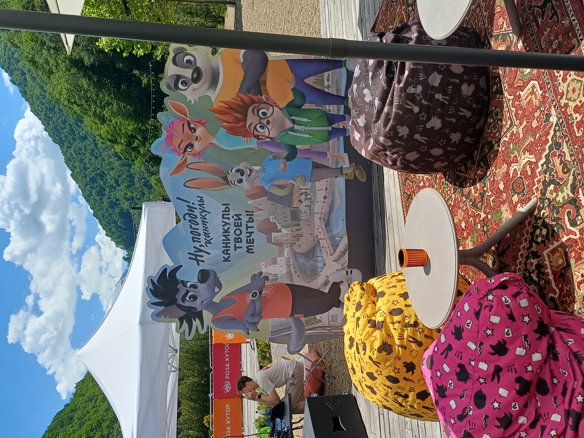 This image captures an outdoor, brightly lit scene at a family-oriented event or recreational area in Estosadok, Russia, likely during the day under a clear sky with scattered white clouds. Lush green, forested mountains dominate the background, emphasizing the natural setting.
In the midground, a large, colorful standee features characters from the Russian animated series "Ну, погоди!" (Well, Just You Wait!), including a wolf, a panda, a rabbit, a deer, and a human-like boy with red hair. The standee's text reads: "Ну, погоди! Каникулы твоей мечты!" (Well, Just You Wait! The Holidays of Your Dreams!). A white canopy tent is pitched behind this standee. To the left, orange banners display "РОЗА ХУТОР" (Rosa Khutor), a prominent resort area, confirming the location context.
The foreground is furnished with several casual seating arrangements. There are vibrant beanbag chairs in yellow, pink, and dark brown, adorned with cartoon character silhouettes and symbols. These are placed around small, round white tables. The ground is covered by a decorative, patterned red and green carpet, adding to a cozy, inviting atmosphere. A man in a light t-shirt and trousers is seated on a folding chair near a black sound speaker, appearing to operate some equipment. The scene suggests a relaxed, family-friendly event or lounge area at a mountain resort.