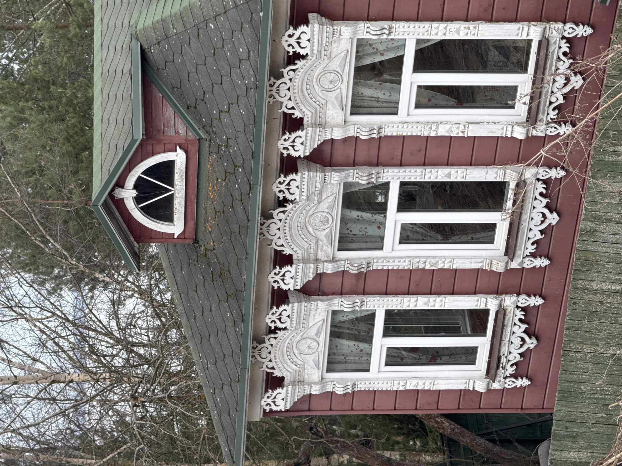 The image depicts a close-up view of a building, specifically its exterior facade. The building appears to be a house or other residential structure.
Architectural features include three vertically aligned windows with elaborate, white-painted carved wooden frames. The frames are ornate, with intricate designs that are a notable architectural element. Above the windows is a roof section with an arched window, also with decorative framing.
The exterior of the building is painted in a reddish-brown color. Some sections of the facade show signs of weathering or age, suggesting the building is not new. Bare tree branches are visible, implying it's likely autumn or winter.
While the prompt mentions Ново-Никольское, Russia, this is not directly apparent from the visual information within the photograph itself. The architectural style is consistent with the location.