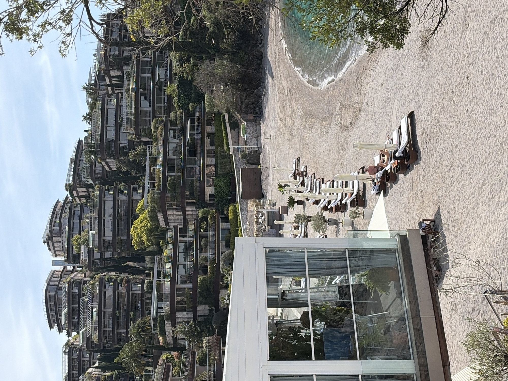 The image depicts a beach resort setting with modern architectural elements, possibly located in or near Budva, Montenegro. The background shows a multi-story building with a curved design and green vegetation integrated into its terraces. In the foreground, there's a beach with lounge chairs occupied by individuals, suggesting relaxation and leisure. A modern, glass-walled structure stands prominently, hinting at a restaurant or recreational area. The beach meets the ocean with gentle waves, reflecting a tranquil coastal environment. The overall scene conveys a luxury or high-end tourist destination.
