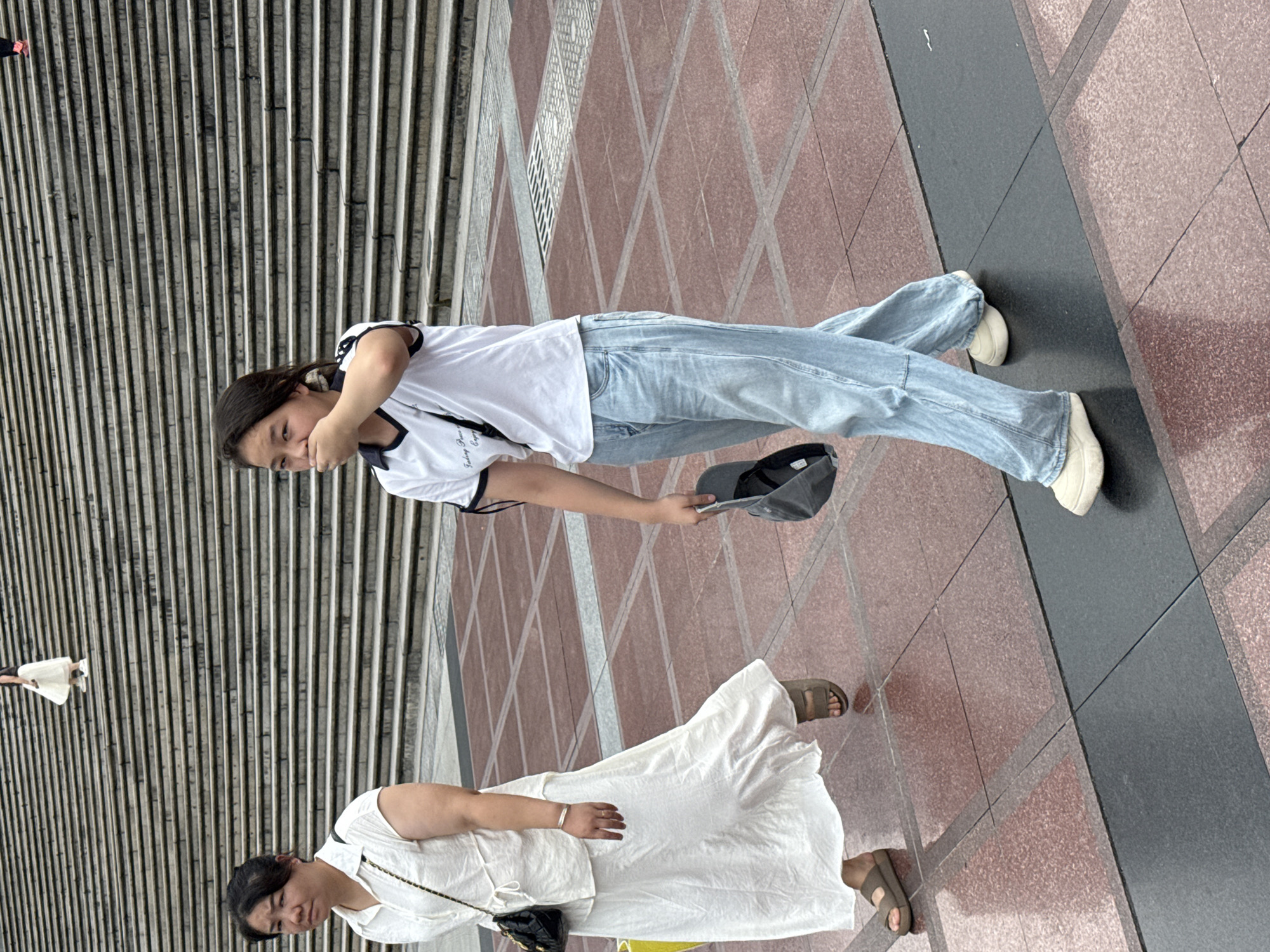 In the Yuzhong District of China, two women are walking on a tiled plaza in front of a wide set of stone stairs. The woman on the left, wearing a white dress and sandals, carries a black purse. The woman on the right, dressed in a white t-shirt and light blue jeans, is holding a gray cap and has her hand to her mouth. The plaza features a repeating pattern of red and black tiles. In the background, another person in a white skirt and black top is ascending the stairs.