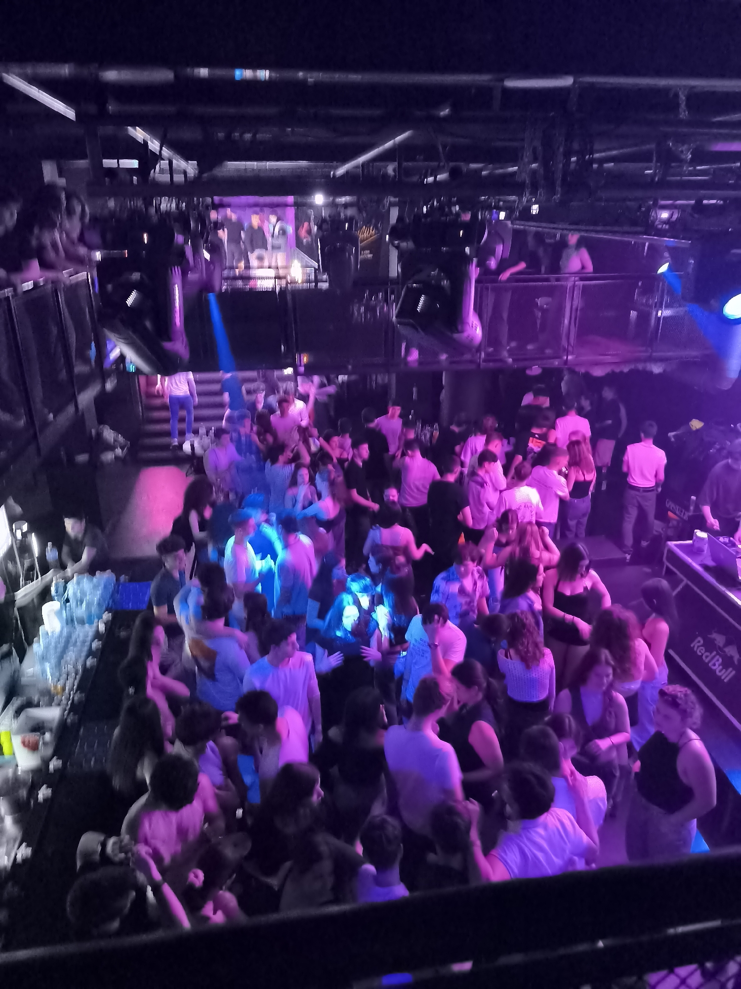 This media file captures a lively, multi-level indoor scene, likely a nightclub or music venue, filled with a large crowd of young adults. The time of day is clearly night, indicated by the dark surroundings and the prominent use of artificial, vibrant lighting, primarily in shades of purple and blue, with some pink and red accents.
The main floor is densely packed with people who appear to be dancing, mingling, and standing around. Their movements, combined with the low light conditions, contribute to a dynamic and somewhat blurry effect throughout the image. To the left, a long bar counter is visible, lined with bottles and cups, where some individuals are gathered. On the right side of the frame, there is a DJ booth, which prominently displays the "Red Bull" logo.
Above the main floor, several upper levels or balconies are visible, also occupied by people observing the scene below or socializing. In the distance, at the back of the venue, there is a raised stage or large screen displaying what appears to be figures, possibly performers or a projected video. Large lighting fixtures and sound equipment are integrated into the venue's structure, enhancing the concert-like atmosphere.
The overall mood is energetic and social, with people enjoying the event. The lighting creates a distinct club aesthetic.