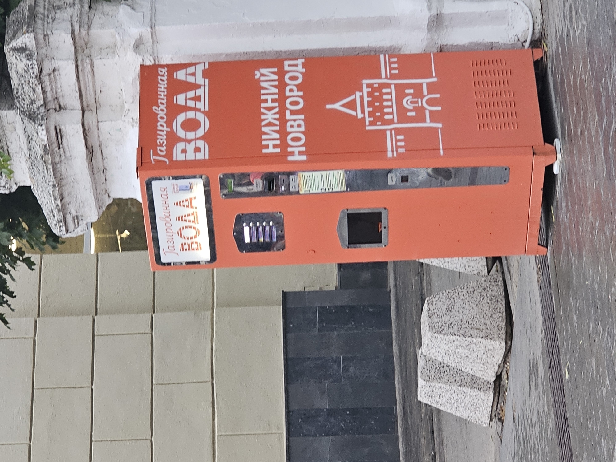 A bright orange vending machine, labeled "газированная вода" (carbonated water) and "Нижний Новгород" (Nizhny Novgorod), stands on a wet, paved sidewalk. The machine has a digital display, a payment slot, and a dispenser. It is adorned with a white graphic of a fortified tower, likely a landmark of Nizhny Novgorod. Behind the machine is a textured, light-colored building facade, and to the right, a white, fluted column. In the foreground, two large, conical concrete bollards with a speckled texture are visible, and a metal grate runs along the edge of the sidewalk. The scene suggests an urban outdoor setting in Nizhny Novgorod, Russia, possibly on a cloudy or rainy day given the wet pavement. There are no people or animals present in the image.