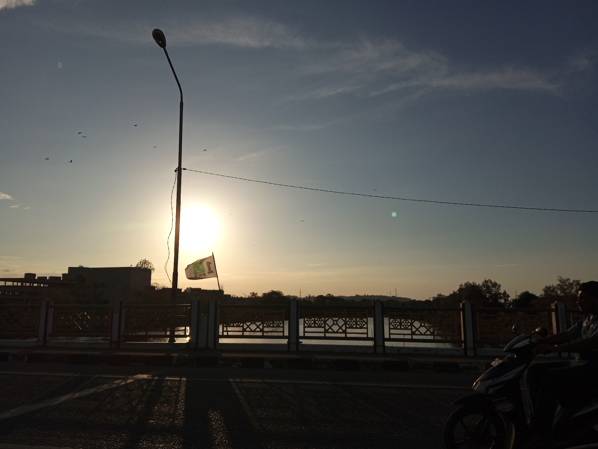 The image depicts a scene in Lhokseumawe, Indonesia, likely on a bridge at sunset. The sun is positioned directly behind a flag attached to a lamp post, creating a silhouette effect. A railing with decorative cutouts runs along the bridge. A person on a scooter is visible on the right side of the frame. Several birds are in flight in the sky. The lighting suggests either sunrise or sunset.